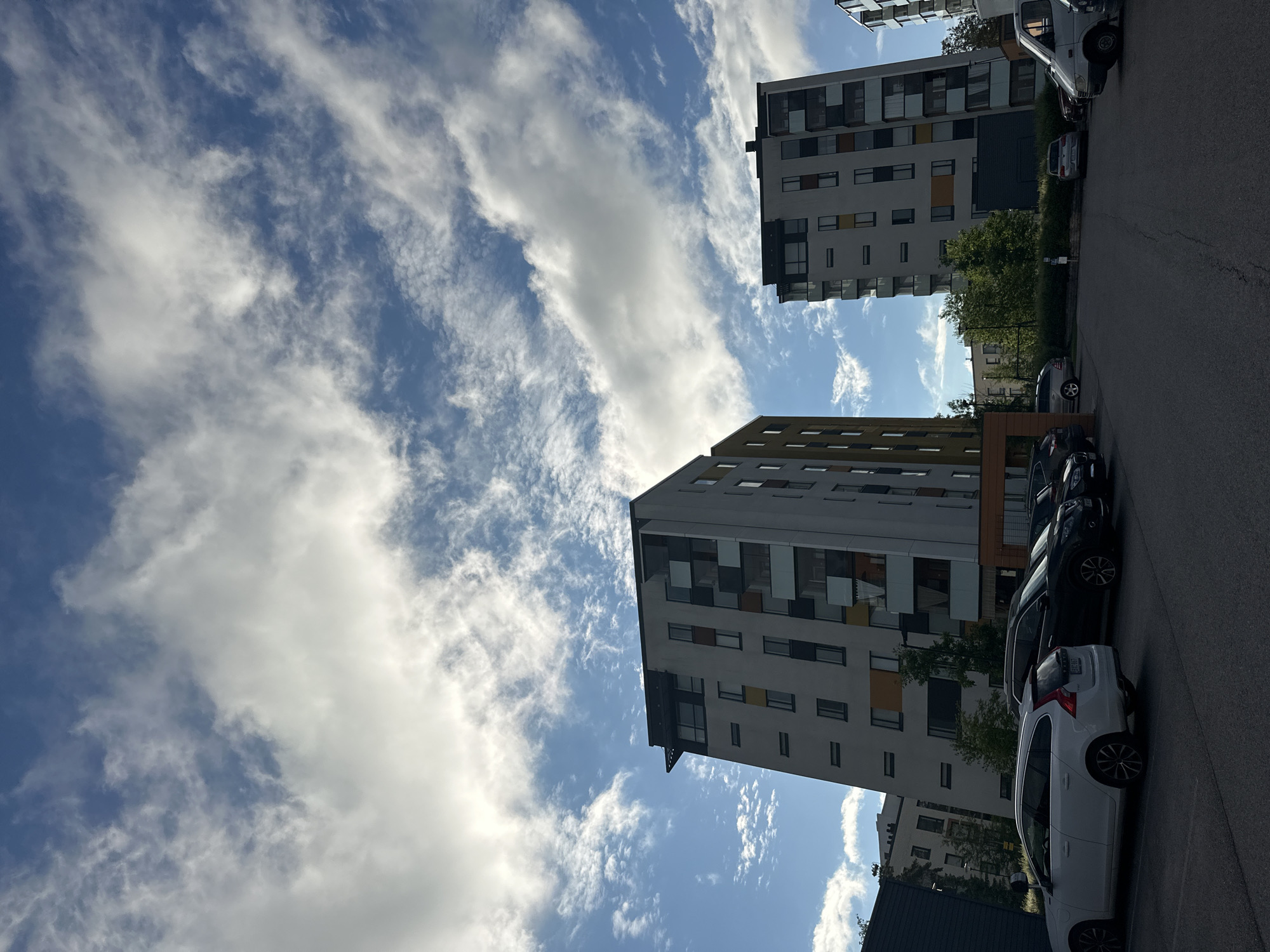 In an outdoor urban scene, two multi-story apartment buildings dominate the frame against a bright blue sky filled with wispy white clouds. The building on the left is a light-colored structure with alternating bands of yellow and dark colored windows, while the building on the right is a taller, predominantly white building with colorful accents around its balconies and windows. Both buildings have a modern architectural style. In the foreground, a parking lot is visible with several cars parked, including a white Volvo station wagon, a dark SUV, and other vehicles. Trees and green foliage are interspersed between the buildings and in the background. The lighting suggests it is daytime, possibly late afternoon, with some shadows cast on the ground. There are no people visible in the scene.