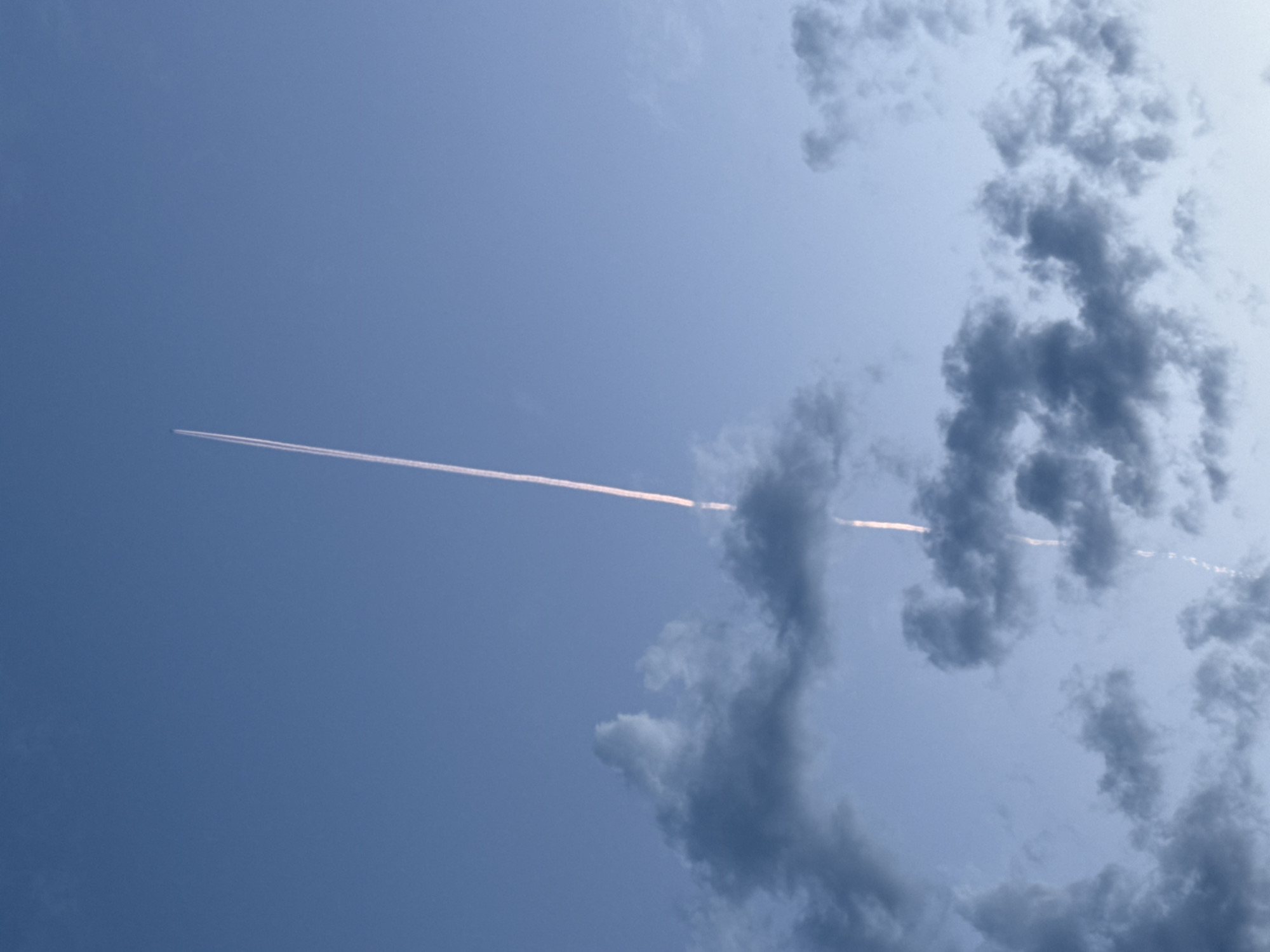 The image shows a blue sky with cumulus clouds on the right side. A contrail is visible stretching across the sky from the upper left to the lower right, partially obscured by the clouds. The sky is bright, suggesting it is daytime. The location, Chaikovsky, Russia, is not apparent from the visual information in the image.