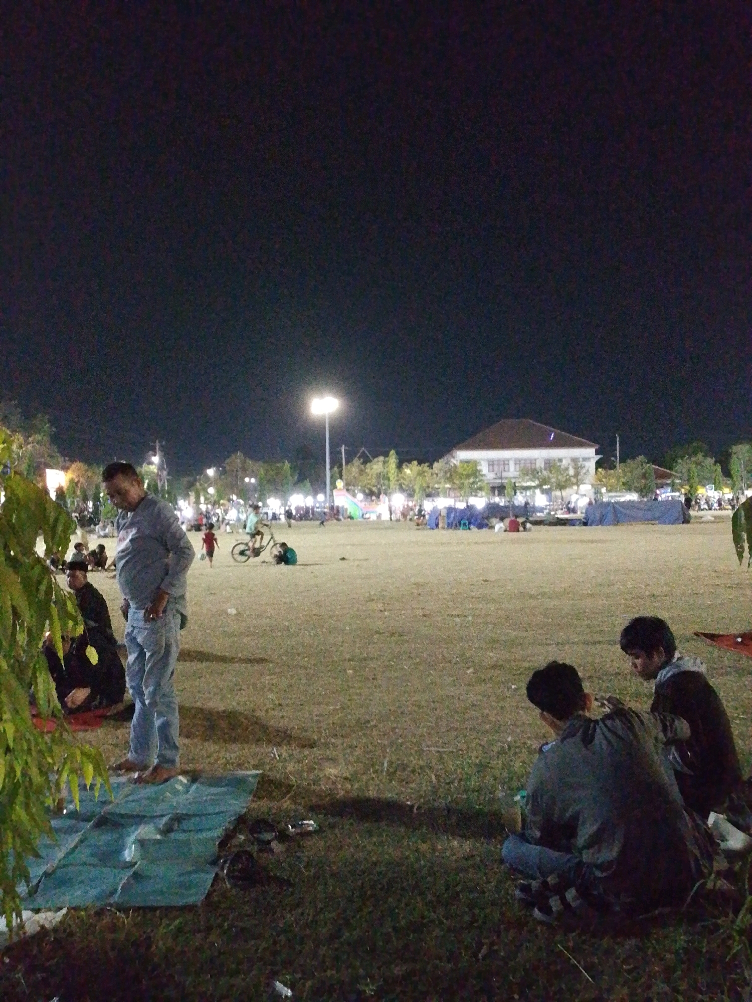 This is a nighttime photograph taken in Tegal, Indonesia, likely at a public open space such as a park or field. The foreground is dominated by a grassy area, with a large, green tarp laid out on the ground in the lower left quadrant. Several people are visible, some sitting and interacting.
In the mid-ground, a man wearing a light blue collared shirt and light blue pants stands and looks downwards. To his left, another person in dark clothing is seated on the ground. Further into the scene, other individuals are scattered across the field. A child in a red shirt is walking, and a person on a bicycle is visible in the background.
In the distance, a long, light-colored building with a brown roof is illuminated, suggesting it might be a community center, school, or government building. Surrounding the building are trees and what appear to be tents or canopies set up on the ground. Streetlights illuminate the open field, casting a warm glow on the scene. The overall atmosphere suggests a relaxed evening gathering or event.