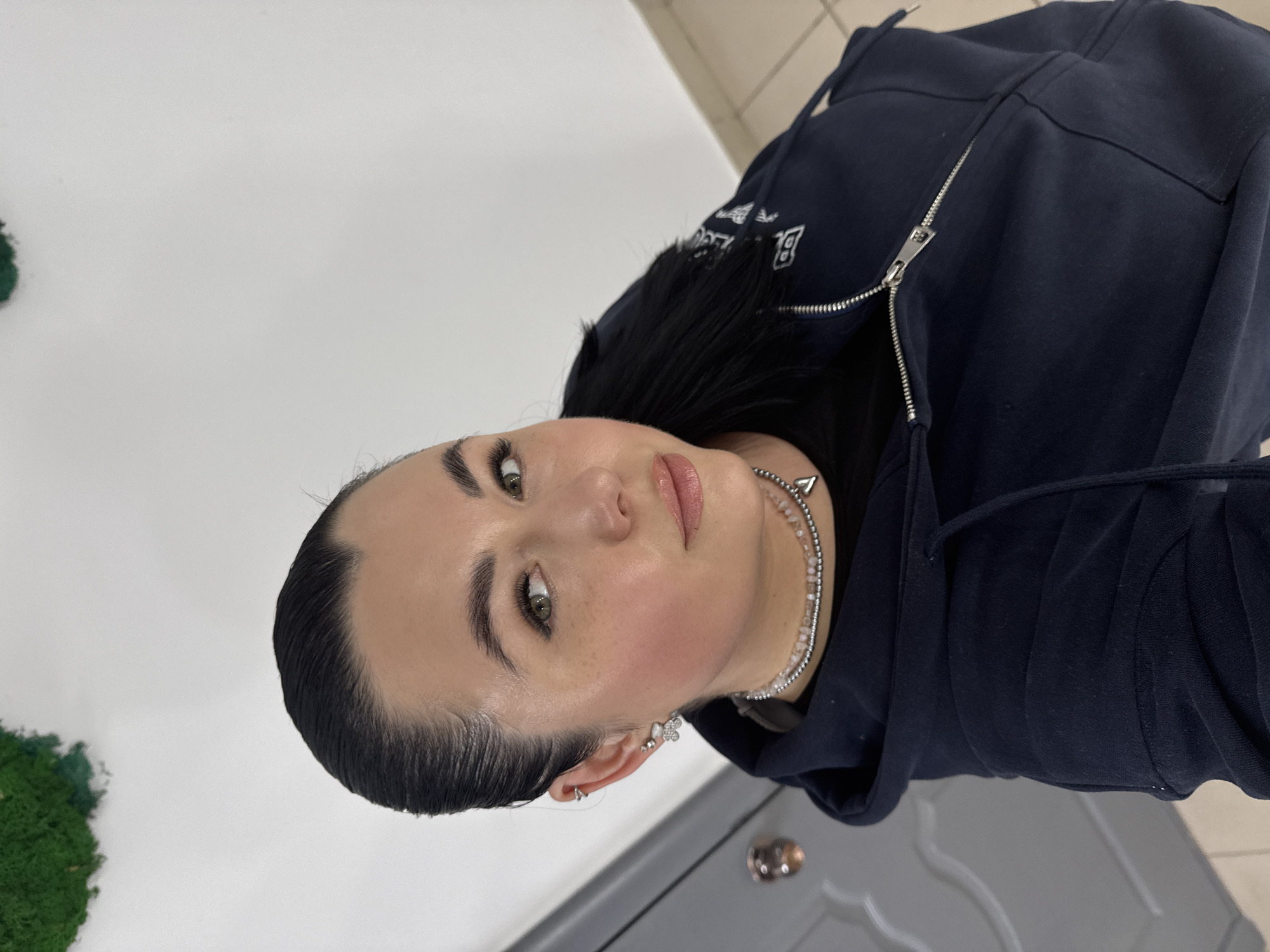 A woman is shown in an indoor setting. She has dark hair styled back from her face, light skin, and green eyes. She is wearing a dark blue zippered hoodie and a beaded necklace. Her face is close to the camera, offering a clear view of her features. The background is a combination of a white wall and what appears to be a gray countertop or cabinet surface. The location of this image is Chaikovsky, Russia.