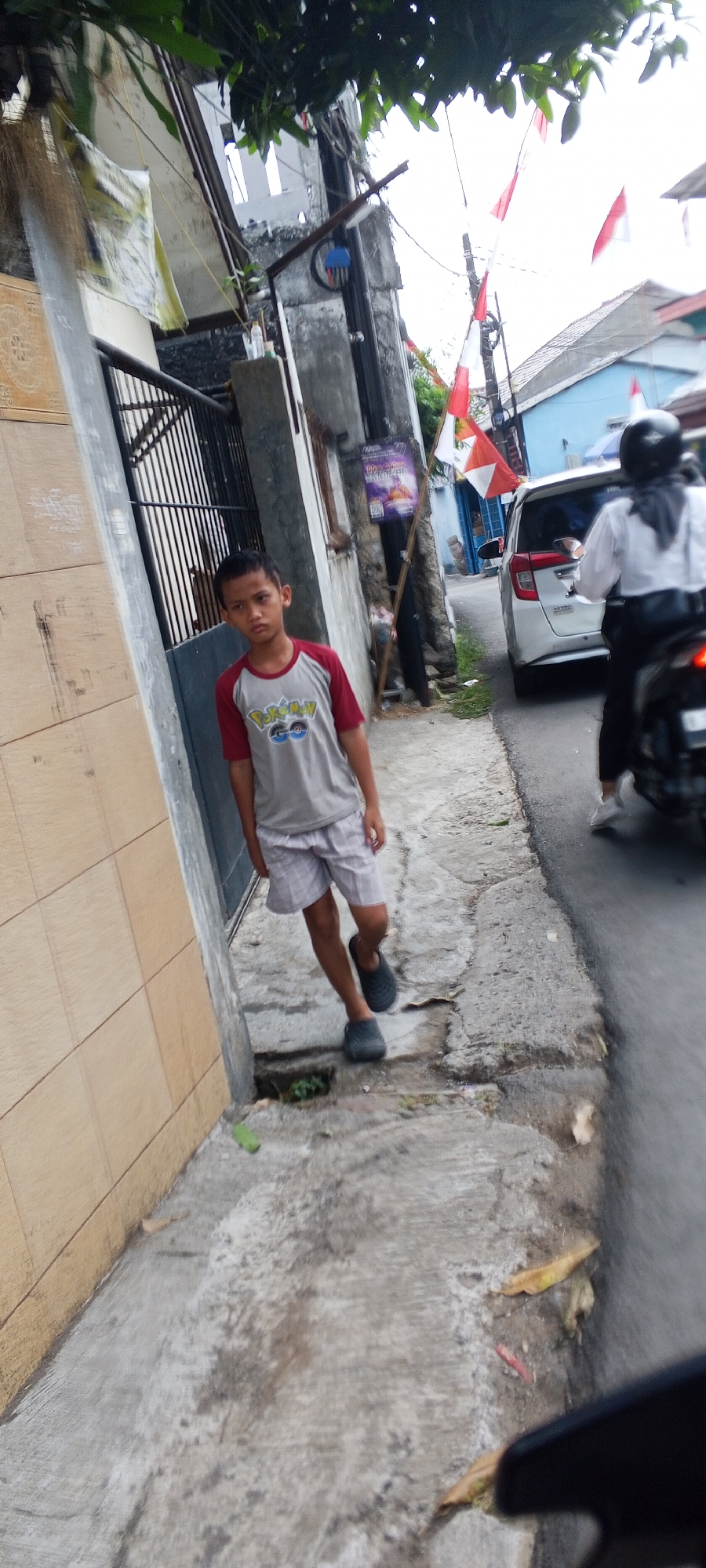 The image depicts a street scene in Depok, Indonesia, with a young boy standing on a sidewalk. The boy, wearing a gray t-shirt with a Pokemon Go logo and light-colored shorts, is looking directly at the camera with a neutral expression. He is wearing dark clogs on his feet.
The sidewalk appears to be made of concrete and is somewhat uneven, with some cracks and debris. To the left of the boy is a textured wall, possibly of a building, and a black metal fence. Behind the boy, a utility pole is visible, adorned with several Indonesian flags, indicating a possible national celebration or holiday.
In the background, a white car is driving on the street, and a person on a motorcycle wearing a helmet and dark clothing is also present. Buildings with tiled roofs are visible further down the street, and lush green foliage can be seen in the upper part of the image, suggesting a tropical environment. The lighting suggests it is daytime, possibly overcast.