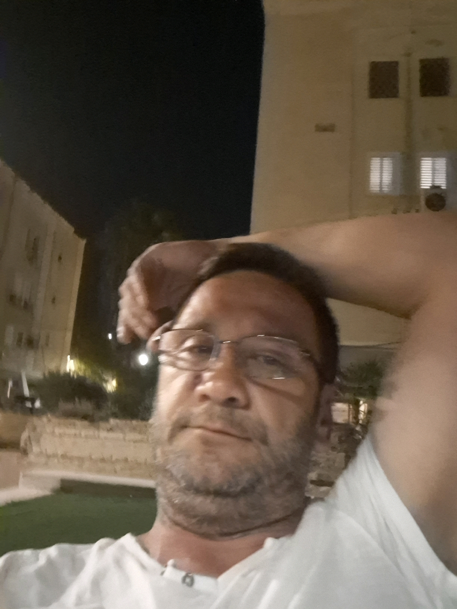 In Yerocham, Israel, at night, a man in a white t-shirt is taking a selfie outdoors. He is wearing glasses and has a beard. His right arm is raised, resting on his head. In the background, a building with lit windows and air conditioning units is visible, along with some greenery and streetlights.
