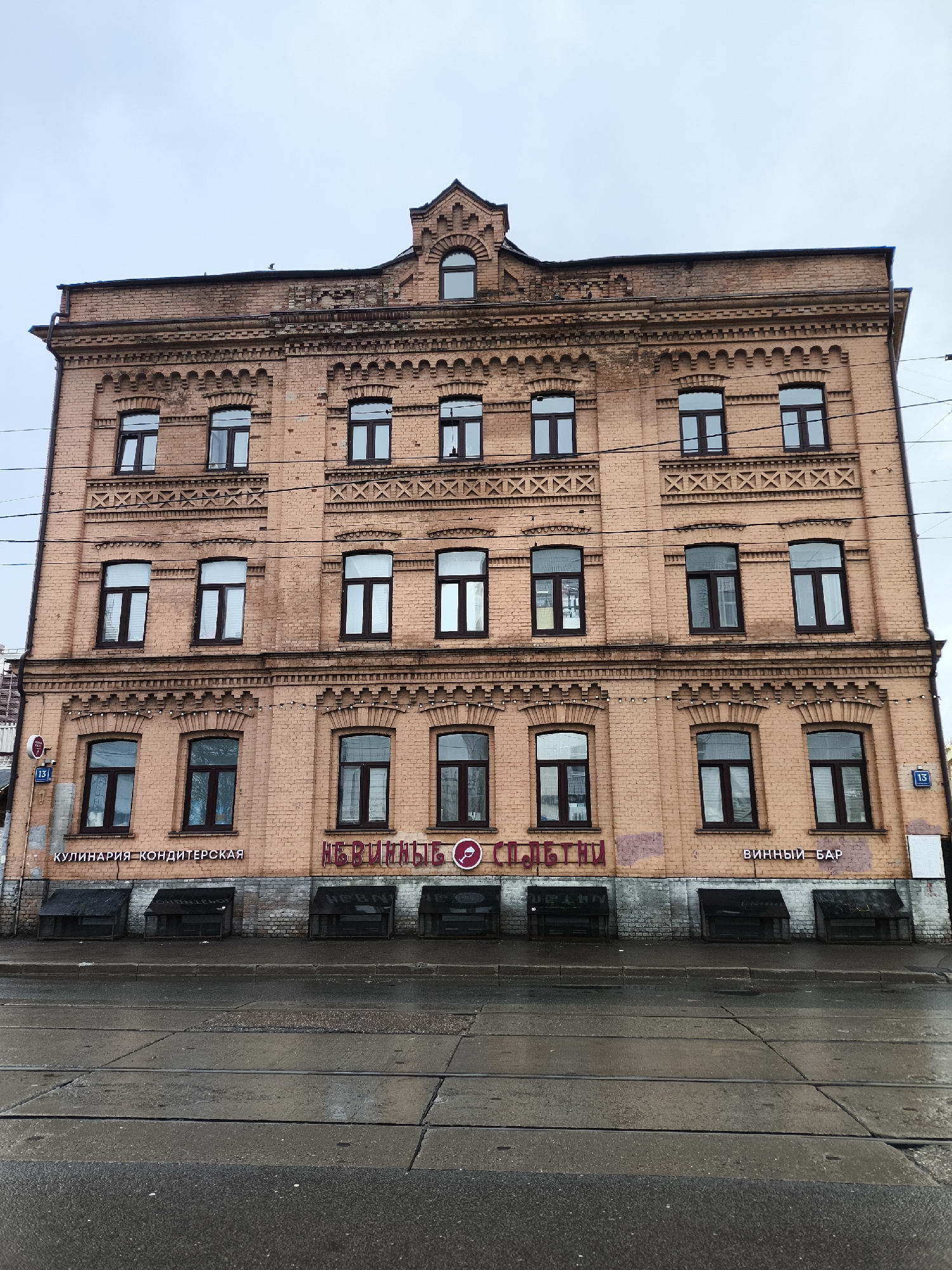 The image displays a four-story brick building with an attic dormer, likely located in Moscow, Russia. The building is constructed from light reddish-brown brick and features ornate architectural details, including decorative cornices, dentil courses, and repeating "X" patterns between the windows of the upper floors. All windows are dark-framed with multiple panes. A distinctive central gable with an arched window is prominent at the roofline.
The scene depicts an overcast day, with a uniformly gray sky. The street and sidewalk in the foreground appear wet, indicating recent rain or damp weather. No people are visible, lending a quiet atmosphere to the urban setting.
At street level, the building houses several businesses. From left to right, visible text includes:
* "КУЛИНАРИЯ-КОНДИТЕРСКАЯ" (Culinary-Confectionery)
* "НЕВИННЫЕ СПЛЕТНИ" (Innocent Gossip), accompanied by a wine glass icon.
* "ВИННЫЙ БАР" (Wine Bar)
Below these storefronts, several dark, enclosed window wells or basement covers line the base of the building. Two blue street signs displaying the number "13" are visible on either side of the lower facade. Overhead power lines crisscross in front of the building. In the immediate foreground, concrete slabs with embedded metal rails for tram tracks are clearly visible, confirming a city location. The building exhibits some signs of age and weathering, yet retains its architectural character.