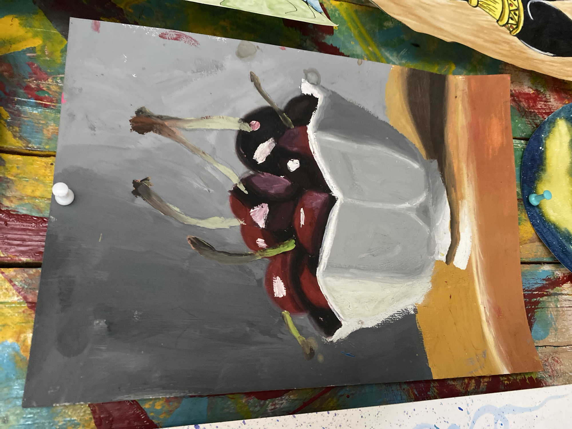The image is a painting of cherries in a bowl. The painting is on a piece of paper that is pinned to a colorful, wooden surface. The cherries are dark red and are depicted with highlights. They are in a light gray, faceted bowl. The background of the painting is a mottled gray. The wooden surface behind the painting is a collage of various colors and textures, suggesting it might be an artist's workspace or a display area. The painting is oriented vertically. There are no people or animals visible in the image. The scene does not depict any specific location in Da Nang, Vietnam.