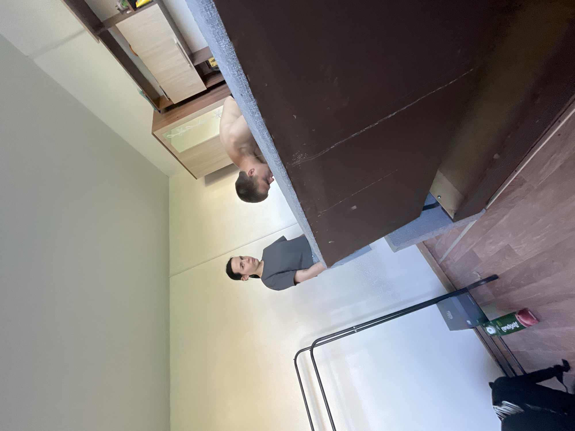 The image depicts an indoor scene, likely a bedroom, with two individuals. A woman with dark hair pulled back into a ponytail stands on the left, wearing a dark grey t-shirt and blue jeans, looking towards the viewer. She is positioned near a black garment rack. On the right, a shirtless man with short brown hair is lying face down on a bed, his head facing away from the camera. The room features a wooden floor, light-colored walls, and a large wooden wardrobe with mirrored panels and shelves on the far right. A juice carton is visible near the garment rack. The lighting suggests daylight. The location is identified as Красные Баки, Russia.