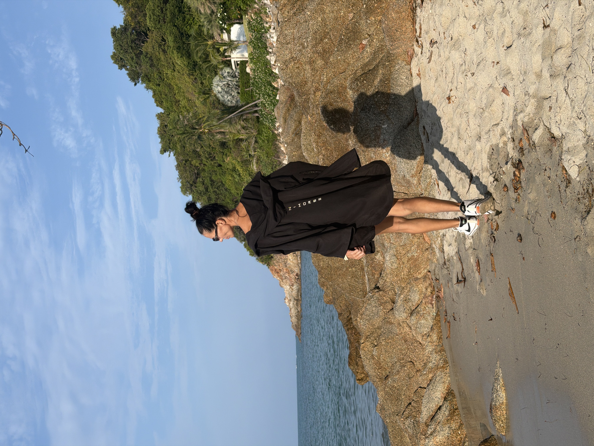 A woman stands on a sandy beach, facing away from the camera and looking towards the ocean. She is wearing a black hooded robe and white sneakers with black accents. The beach is lined with large rocks and the ocean stretches out before her. In the background, a lush green hillside rises, dotted with palm trees and a white building. The sky is a clear blue with some wispy clouds. The scene appears to be a tranquil coastal landscape. The specific location is the Koh Samui District in Thailand.