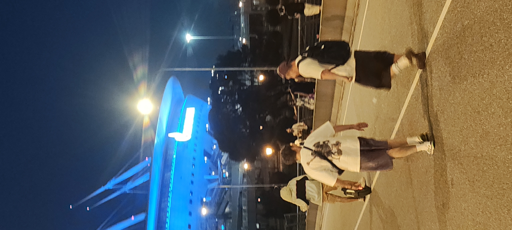 An outdoor, eye-level, slightly upward-angled, vertical, full-color photograph taken at night on an elevated roadway in Saint Petersburg, Russia, depicts several individuals engaged in various activities. The prominent feature in the upper background is a large, modern, blue-lit structure, possibly a stadium or arena, with illuminated supports. A bright, diffused light source, likely a streetlamp or the moon, is visible in the upper right quadrant, casting lens flares.
In the mid-ground, a person on a unicycle rides away from the viewer, wearing a light-colored hooded jacket with a dark horizontal stripe and khaki shorts. To their right and slightly ahead, another person walks away from the viewer, wearing a white t-shirt with a graphic print on the back, dark shorts, and white socks with sandals. This individual is carrying a black bag slung across their chest. Further to the right, a third person walks away from the viewer, wearing a white t-shirt, a dark skirt or kilt, and a backpack, with a maroon baseball cap on their head.
In the background, other individuals are visible on the same roadway, some standing and others walking, appearing to be part of a crowd. Trees with dark foliage are scattered throughout the mid-ground and background. Several streetlights with warm yellow illumination dot the scene. The roadway itself is paved and marked with white lines. The overall atmosphere suggests a public gathering or event occurring in an urban setting at night.