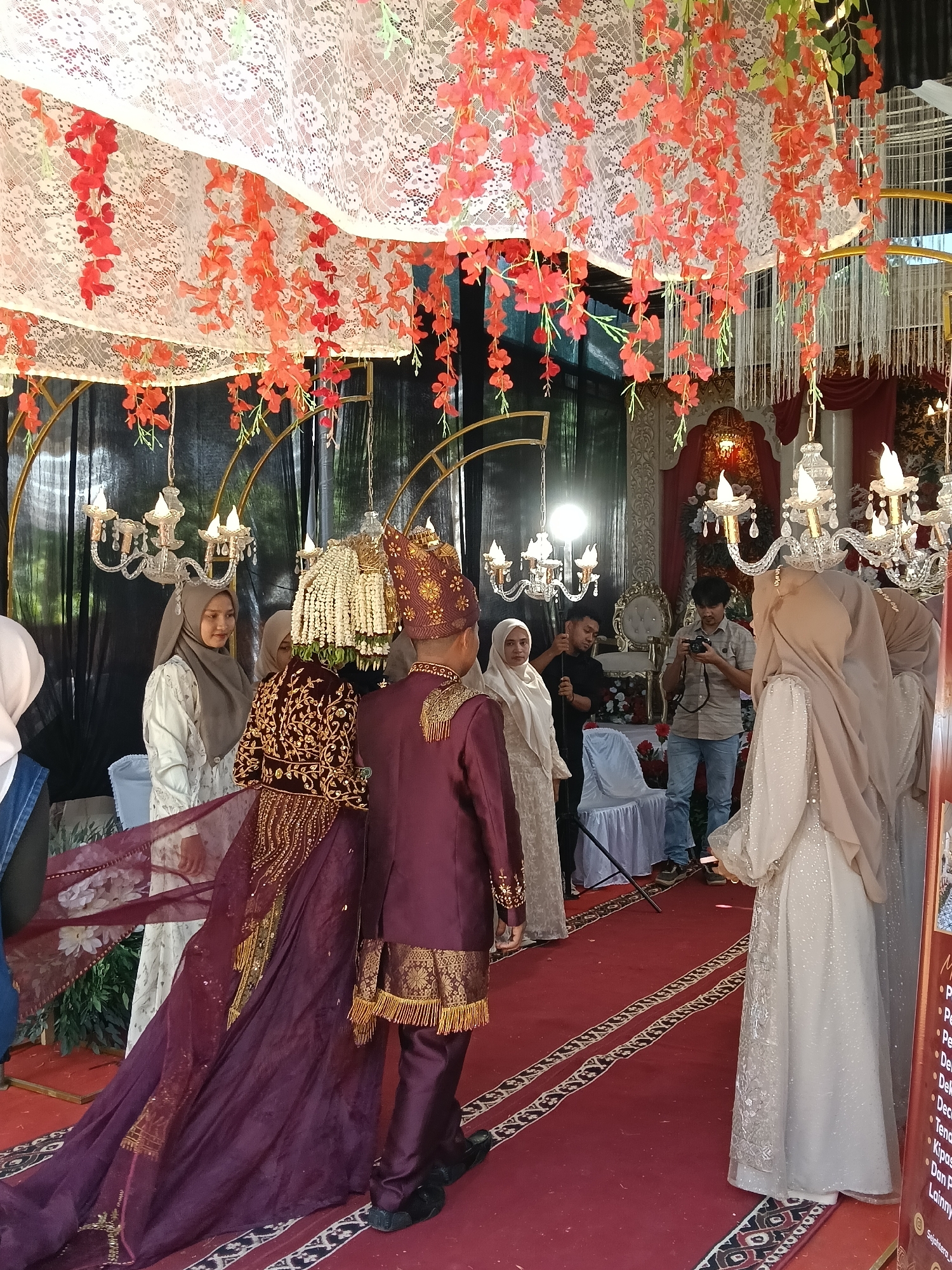 At a wedding ceremony in Geulumpangsulu Timu, Indonesia, a couple stands on a red carpet decorated with geometric patterns. The groom wears a maroon suit with gold embroidery, a matching hat, and traditional beaded headwear, while the bride is adorned in a deep maroon gown with intricate gold embellishments and a flowing train, also wearing traditional headwear. They are surrounded by guests, some of whom are capturing the moment with cameras. The venue is decorated with white lace fabric, cascading red flowers, and ornate chandeliers. A photographer is visible in the background, focusing on the couple. The overall atmosphere suggests a formal and celebratory occasion.