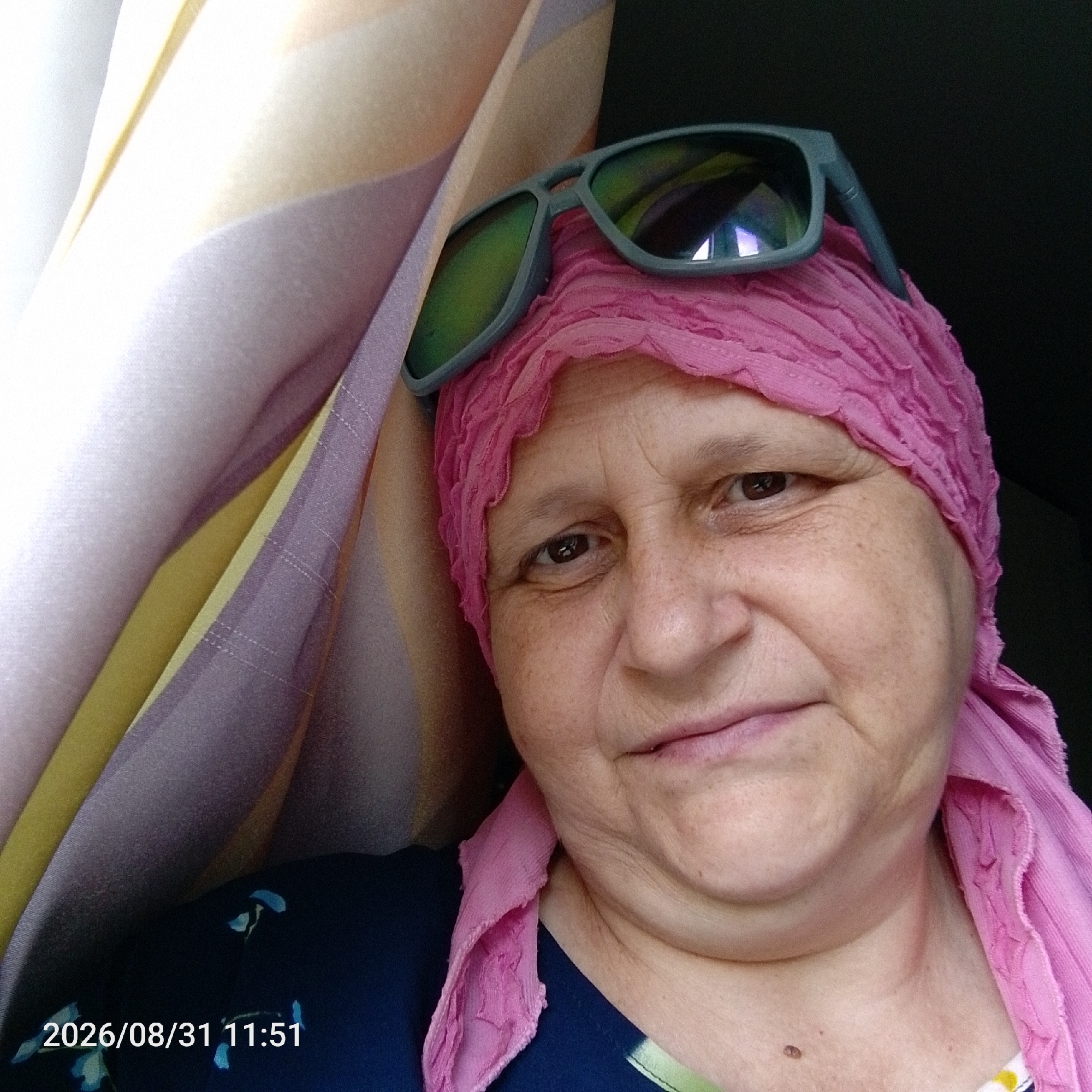 A close-up, selfie-style photograph shows a middle-aged woman with fair skin and freckles, wearing a pink headscarf and sunglasses. The sunglasses are grey with green lenses and are resting on top of her headscarf. She is looking directly at the camera, with her mouth slightly open in a smile. The background is a curtain with a striped pattern in shades of beige, yellow, and purple, and a dark area to the right. A timestamp of "2026/08/31 11:51" is visible in the bottom left corner of the image, superimposed over a dark blue garment with a floral pattern. The lighting suggests an indoor setting, possibly near a window.