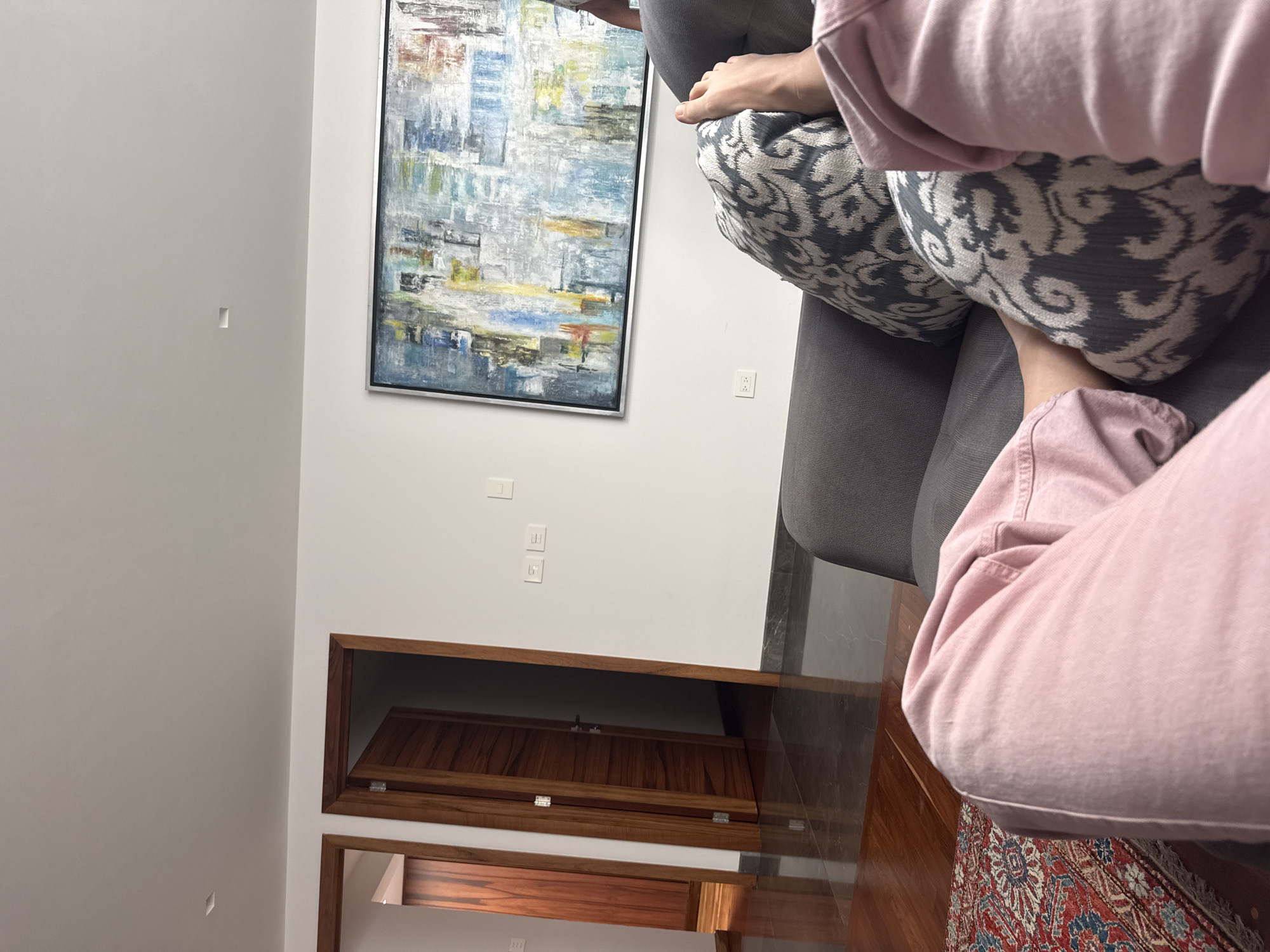 The image shows an interior space with white walls and a dark wood floor. On the wall is an abstract painting with blue, gray, and yellow tones. There are also electrical outlets and a wooden cabinet or door visible on the lower portion of the wall. A person in pink clothing is sitting on a gray sofa with a patterned pillow. The edge of a patterned rug is visible on the floor. The location is presumed to be in San Bernardino Tlaxcalancingo, Mexico, based on the prompt.