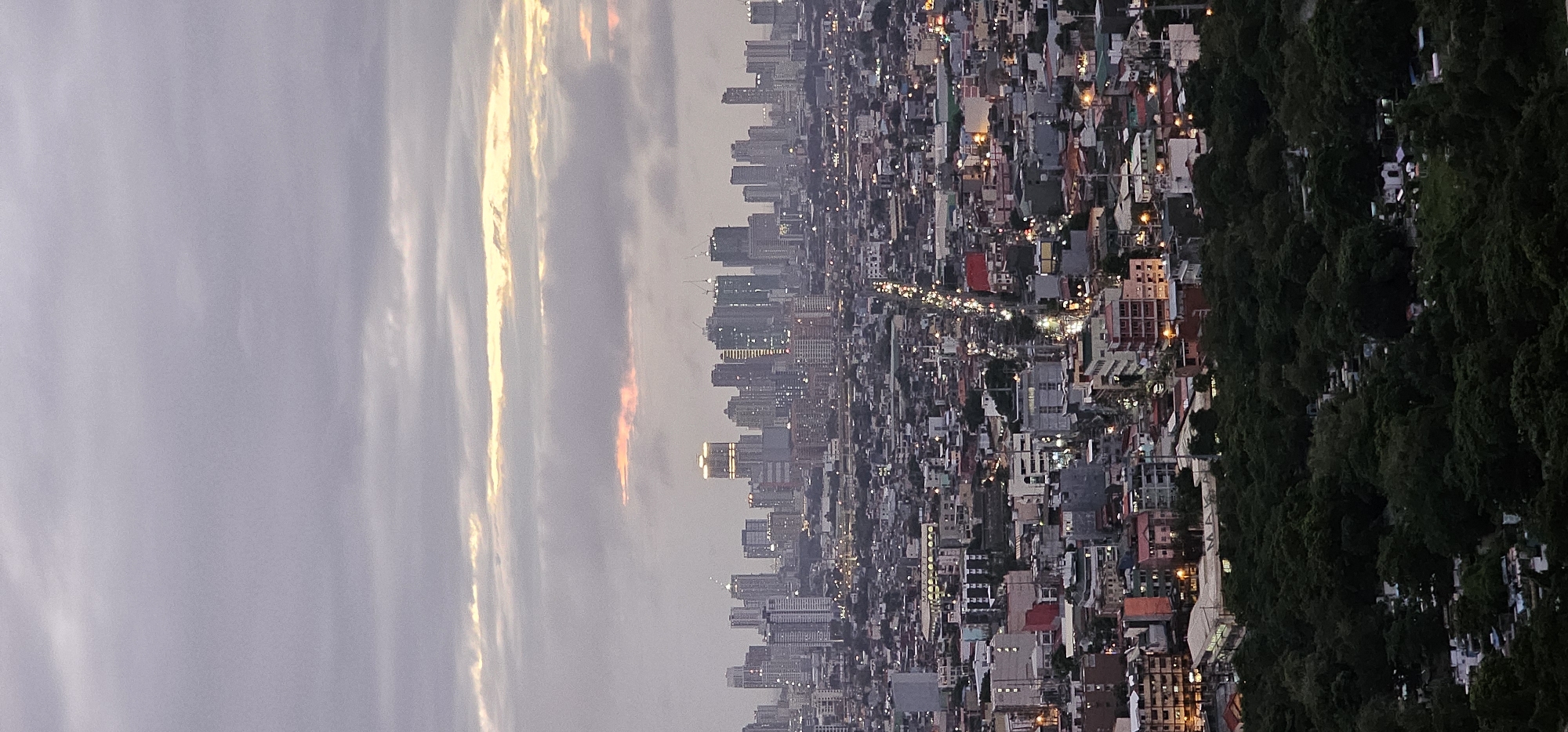This is an aerial, vertical photograph of the Makati skyline in the Philippines at dusk. The sky is overcast with a muted palette of grays and purples, with streaks of pale yellow and pink from the setting sun breaking through the clouds.
The foreground is dominated by a dense, dark green canopy of trees, suggesting a park or a heavily wooded area. Within this greenery, there are visible patches of tombstones, indicating the presence of a cemetery.
Beyond the trees, the mid-ground reveals a dense urban landscape composed of a mix of low-rise buildings and apartment complexes. Many of these structures have illuminated windows, suggesting that people are inside and the city is beginning to light up for the evening. A prominent street runs through this section of the city, illuminated by streetlights and the headlights of vehicles, indicating ongoing traffic.
The background showcases the iconic Makati skyline, characterized by a collection of modern skyscrapers of varying heights and architectural designs. Some of these buildings are illuminated, highlighting their structures against the twilight sky. Construction cranes are visible atop some of the taller buildings, indicating ongoing development. The overall impression is a vast urban expanse transitioning from day to night.
