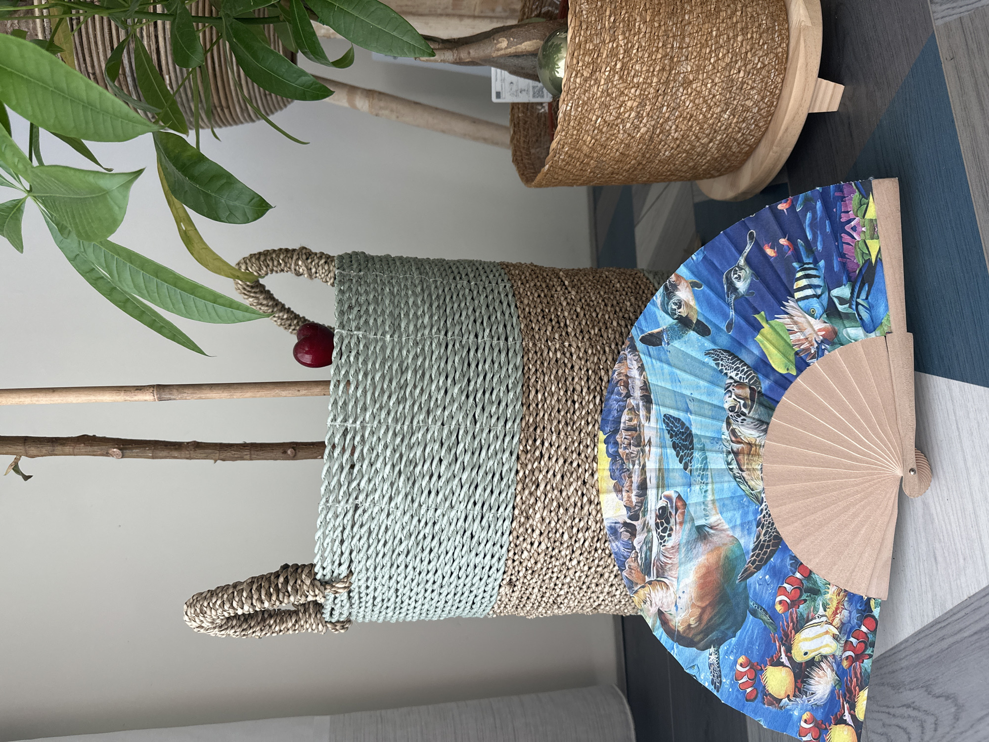 The image shows a close-up of a room interior, with several items arranged on a surface. The foreground features a woven basket, split into two color sections: a light blue at the top and a tan color below. The basket has a handle. Leaning against the basket is a folding fan decorated with images of sea life, including turtles and various fish. A light-colored, possibly wooden, stick is visible inside the basket, topped with a red decorative ball. In the background, two woven plant pots are partially visible. The plant pots are on the floor which appears to be decorated with geometric blue patterns. The room is likely located in Amsterdam, Netherlands.