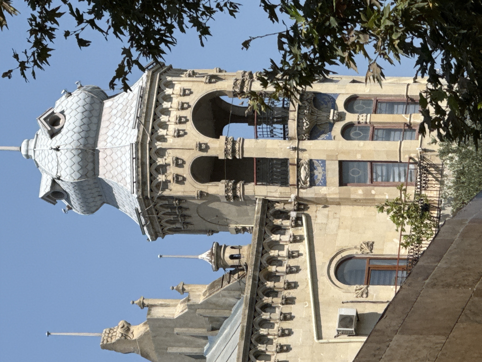 The image shows an exterior view of a historical building in Sabail Raion, Azerbaijan. The building features ornate architecture with a prominent tower topped by a domed roof. The tower has multiple levels with arched openings and decorative elements, including what appear to be sculpted faces. Below the tower, there is a building section with a decorative frieze composed of repeated archways and sculpted figures. Several windows with wooden frames are visible on this section. An air conditioning unit is mounted on the wall of a lower section of the building. A metal balcony with intricate railing overlooks a plant on the balcony. In the foreground and right side of the image, there are large tree branches with dark green leaves, partially obscuring the building. The sky is clear blue. The overall impression is of an old, grand structure, possibly a palace or a historic administrative building, set against a bright sky.