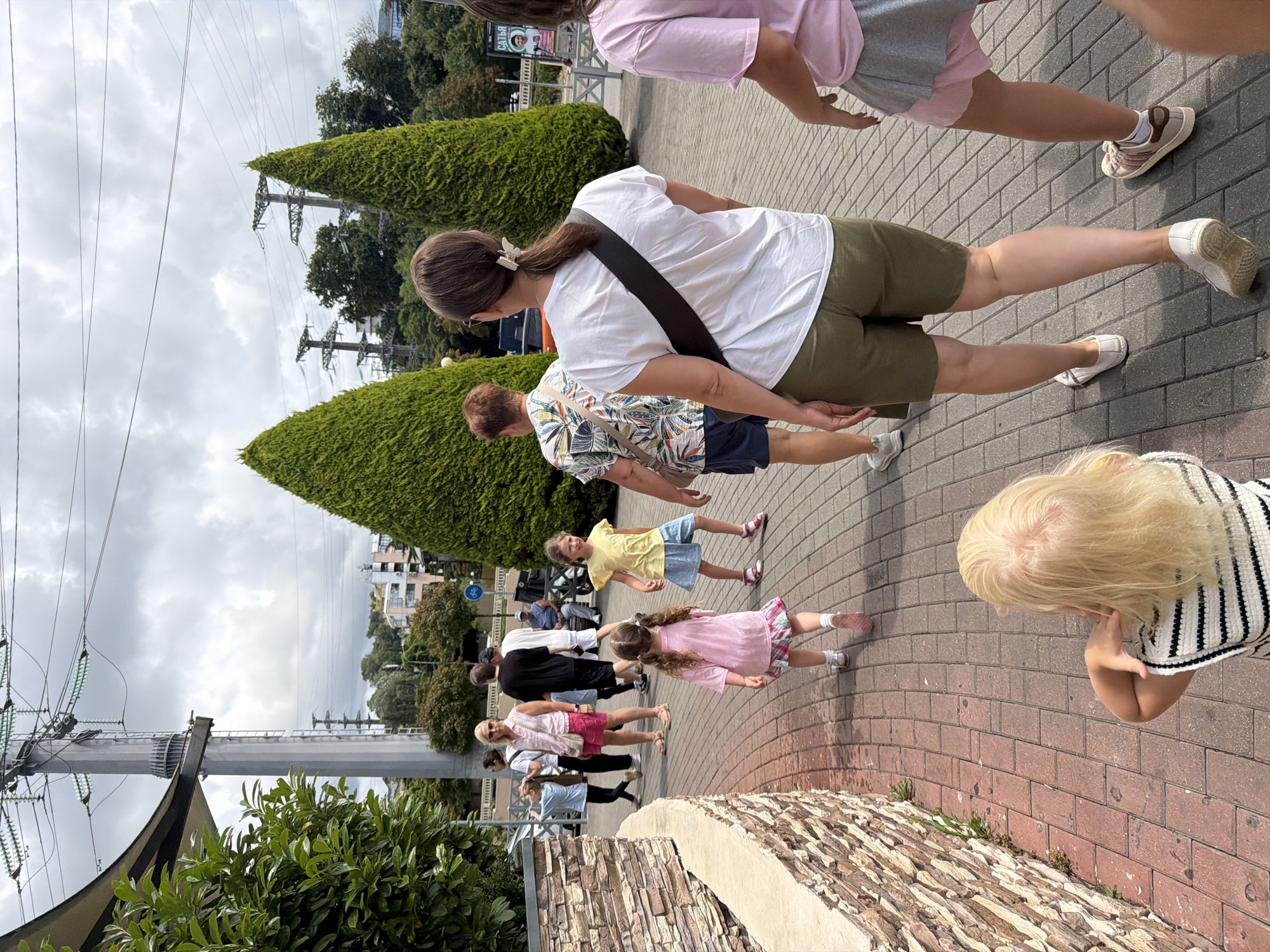 The image depicts a family walking along a paved walkway in Sochi, Russia, near the Olympic Park. The walkway is made of red brick pavers, bordered by a low stone wall on the left. In the background, towering green conical trees flank the path. Overhead, power lines and a large electricity pylon dominate the sky, suggesting proximity to infrastructure.
Several individuals are visible in the frame, all facing away from the camera and moving in the same direction. In the foreground, a young girl with blonde hair and a striped top is seen from behind. Slightly ahead of her, a young girl in a pink t-shirt and a patterned skirt walks. Further back, a woman in a white t-shirt and olive green shorts, with a dark strap across her chest, leads the way. To her left, a woman in a floral print shirt and blue shorts is also walking. Other figures, including children and adults, are further down the path, their details less distinct. The overall atmosphere suggests a casual stroll in a public outdoor space.