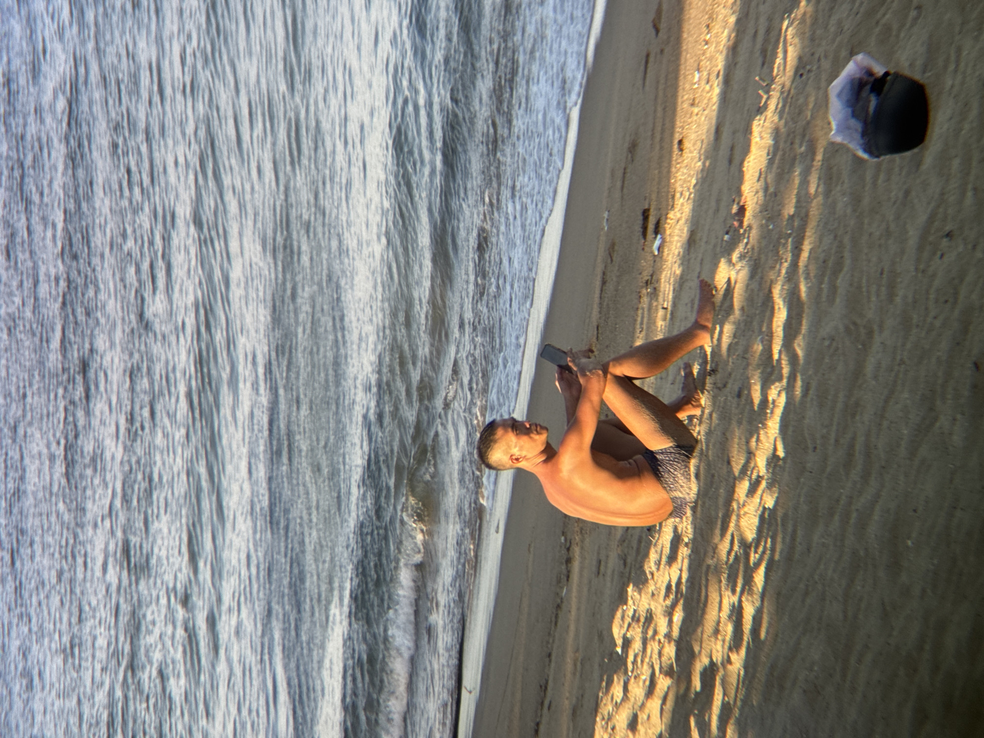 A shirtless man wearing patterned swim trunks sits on a sandy beach in Nha Trang, Vietnam, looking at a smartphone. The man is positioned in the mid-right of the frame, with the ocean occupying the upper half of the image. Waves are breaking gently on the shore. The sand is textured with ripples and shows patches of sunlight and shadow. In the lower right corner, a dark object, possibly a hat, rests on the sand. The overall scene conveys a sense of relaxation and solitude.