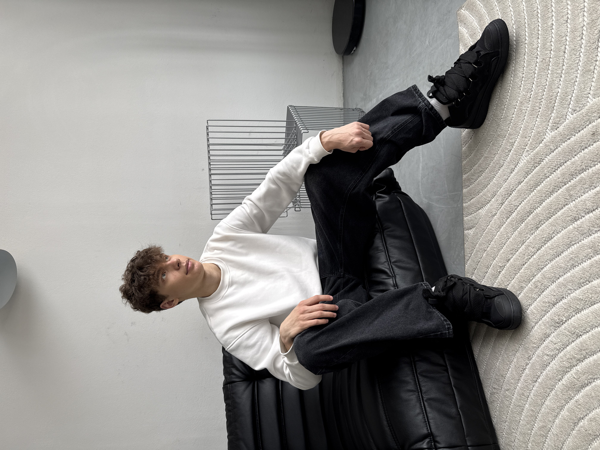 A young man with curly brown hair sits on a black leather couch in what appears to be an indoor setting. He is wearing a white long-sleeved shirt and dark-colored jeans. His feet are clad in black sneakers with prominent laces.
Next to the couch is a wireframe structure, possibly a side table or shelf. The background consists of a white wall and a portion of a grey, textured floor covering, potentially a rug. There is also a black circular object mounted on the wall in the upper left corner.
No information is available to determine the location of the scene, and the image does not provide data or context to support the assertion that the scene is located in Rusanovo, Russia. The available details are limited to the description of the visible elements in the image.