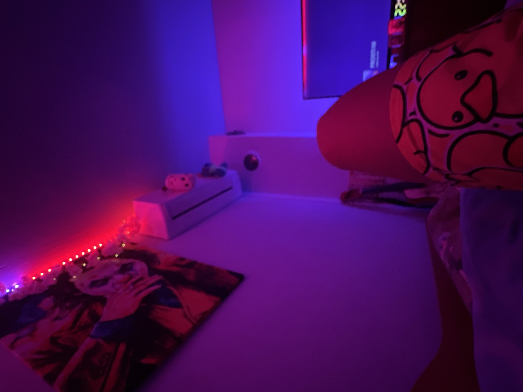 The image depicts an indoor scene, likely a residential room, illuminated predominantly by artificial, colored light, creating a low-light environment. The dominant hues are purple and red on the left side, transitioning to blue on the right. This scene is located in Debelt, Bulgaria.
In the immediate foreground, from a first-person perspective, are the lower extremities of an individual, presumably lying down or reclining. One leg is partially visible, appearing covered by a reddish-brown material or skin, and the knee/lower thigh is covered by a distinct yellow garment or blanket featuring a repetitive pattern of outlined cartoon ducks with orange beaks and small black eyes. The surface beneath the individual is covered by a blue or purple blanket or bedding.
On the upper left wall, a large, rectangular piece of artwork, possibly a canvas or poster, is affixed. It depicts obscured figures or faces in dark and red tones, heavily influenced by the ambient red lighting, making precise details indistinct. Below the artwork, a horizontal string of small, illuminated red LED lights is affixed to the wall.
Further down the left wall, a white wall-mounted split-unit air conditioner is visible. A small, pink, possibly pig-shaped plush toy with distinct black eyes rests atop the air conditioner unit. Below the air conditioner, a circular, dark-colored device with a central lighter circle is mounted on the wall.
On the far right wall, a flat-screen television is displaying content, showing partial text that appears to be "PROGRAM" against a blue background, consistent with the room's blue illumination in that area. Below the television, a digital display shows green numbers, indicating "22:--" (likely 22:XX, signifying time). Other indistinct, colorful objects are arranged beneath the television screen.
