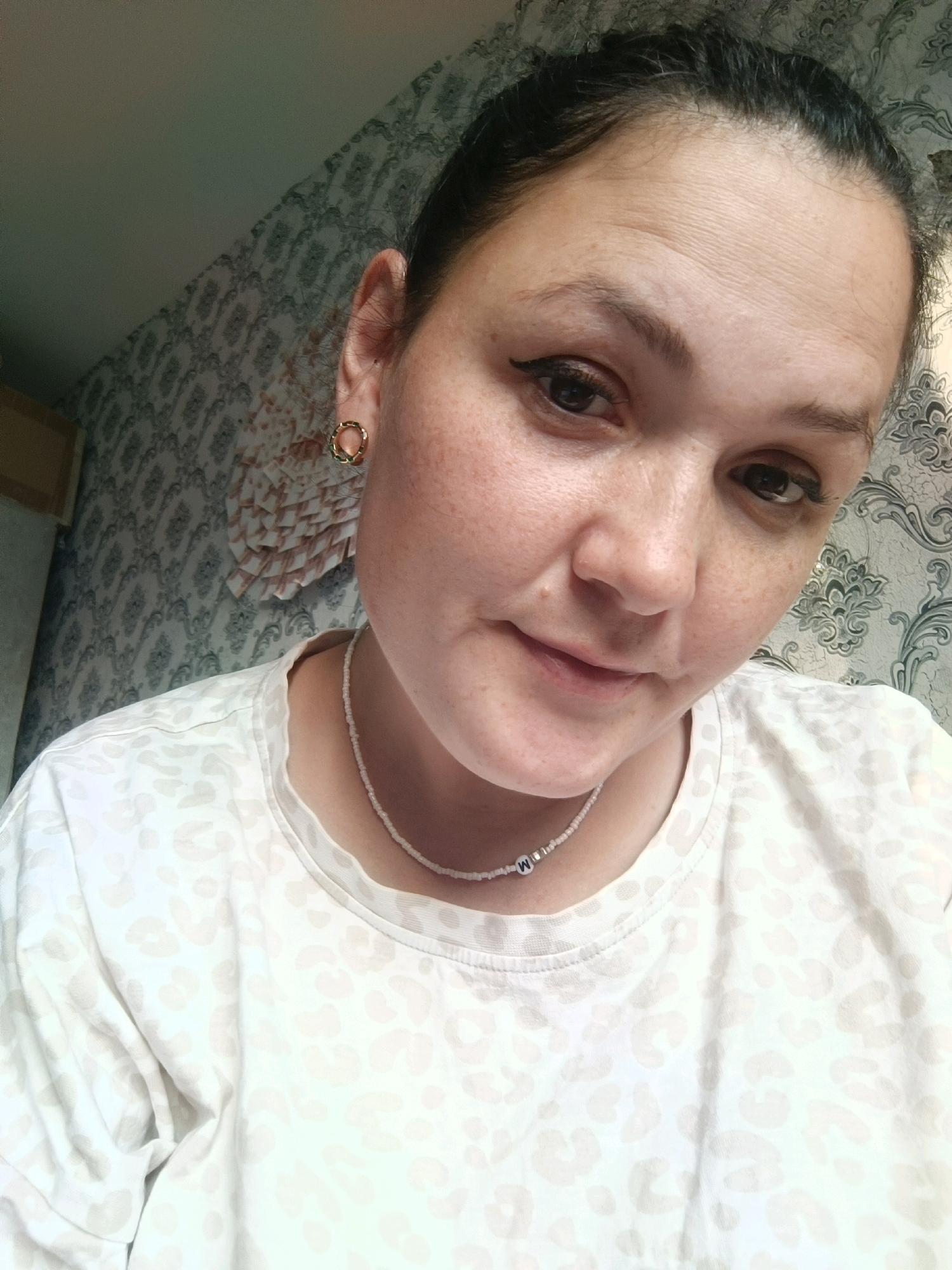 A woman is shown from the chest up, looking into the camera. She has dark hair pulled back, is wearing a light-colored shirt with a leopard print pattern, a beaded necklace, and gold hoop earrings. The background appears to be wallpaper. No specific location in Chaikovsky, Russia, can be identified from the image.