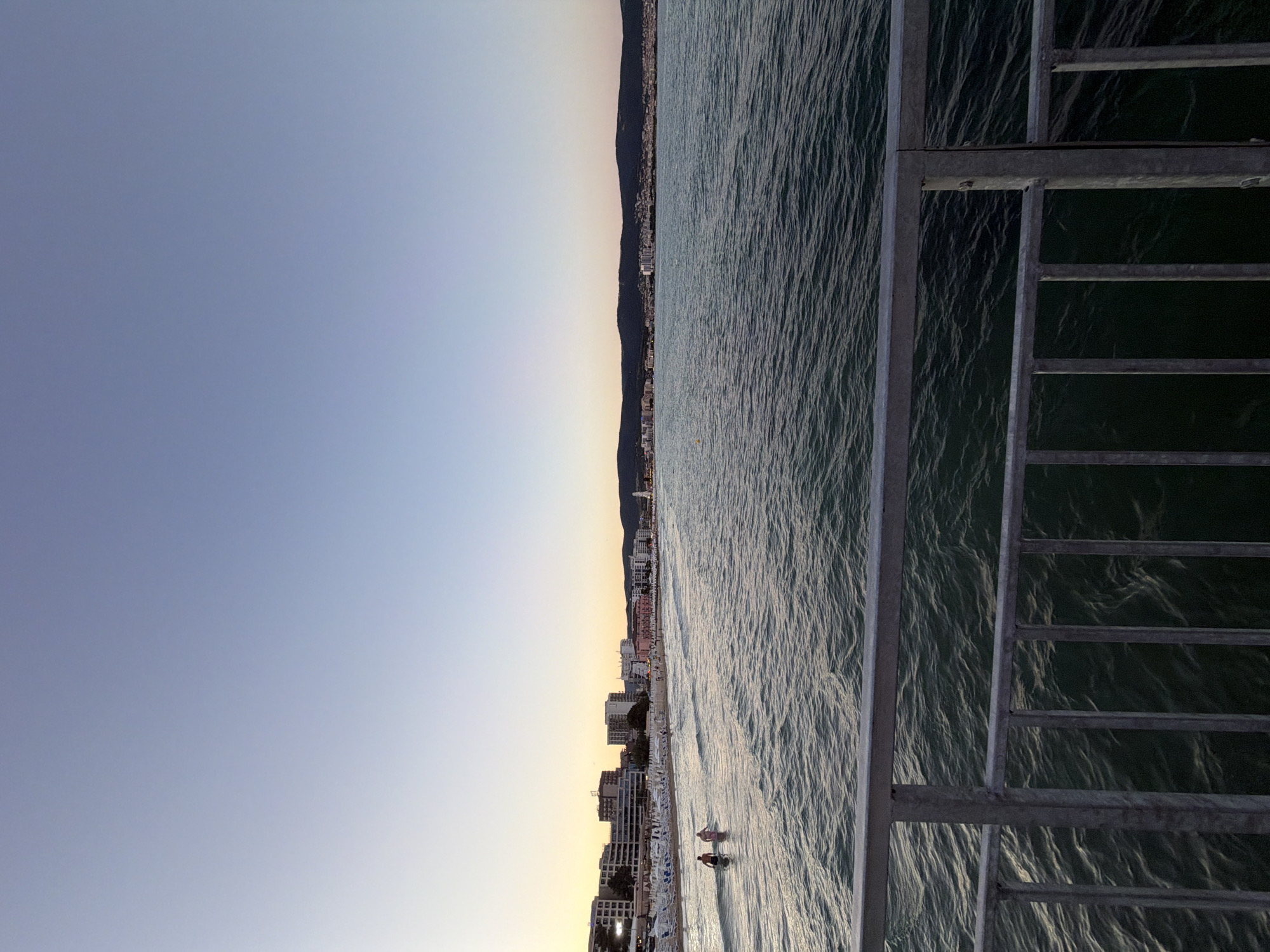 The image depicts a seaside view of Nesebar, Bulgaria, during sunset. The sky transitions from a pale yellow near the horizon to a light blue overhead. In the foreground, a metallic railing with vertical bars overlooks the dark, rippling water. The middle ground features a beach lined with white lounge chairs and umbrellas, behind which are buildings of various heights, some with balconies. A few people are visible on the beach and wading in the shallow water. In the distance, a long, dark mountain range forms the horizon, with a scattering of lights visible along the coastline. The overall scene conveys a sense of a tranquil evening at a popular seaside resort.