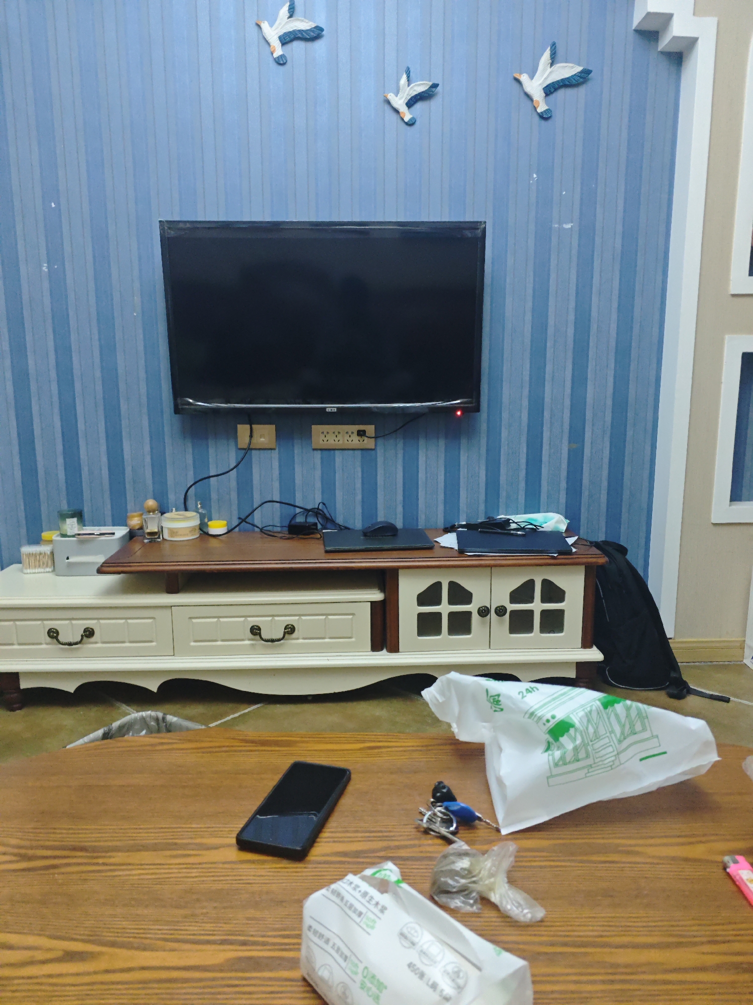 The image shows a room in Chongqing High-tech Zone, China. The scene is centered on a wooden table in the foreground cluttered with personal items. A black smartphone, a set of keys, a clear plastic bag, a pink lighter, and two crumpled white paper bags are scattered across the table's surface. Behind the table, there's a light-colored TV stand with drawers and cabinets, topped by a dark wooden surface. A flat-screen television is mounted on a blue-striped wall above the stand. Three decorative bird figurines are affixed to the wall above the television. A black jacket is draped to the right of the TV stand. The room appears to be a living area or a common space within a residence.