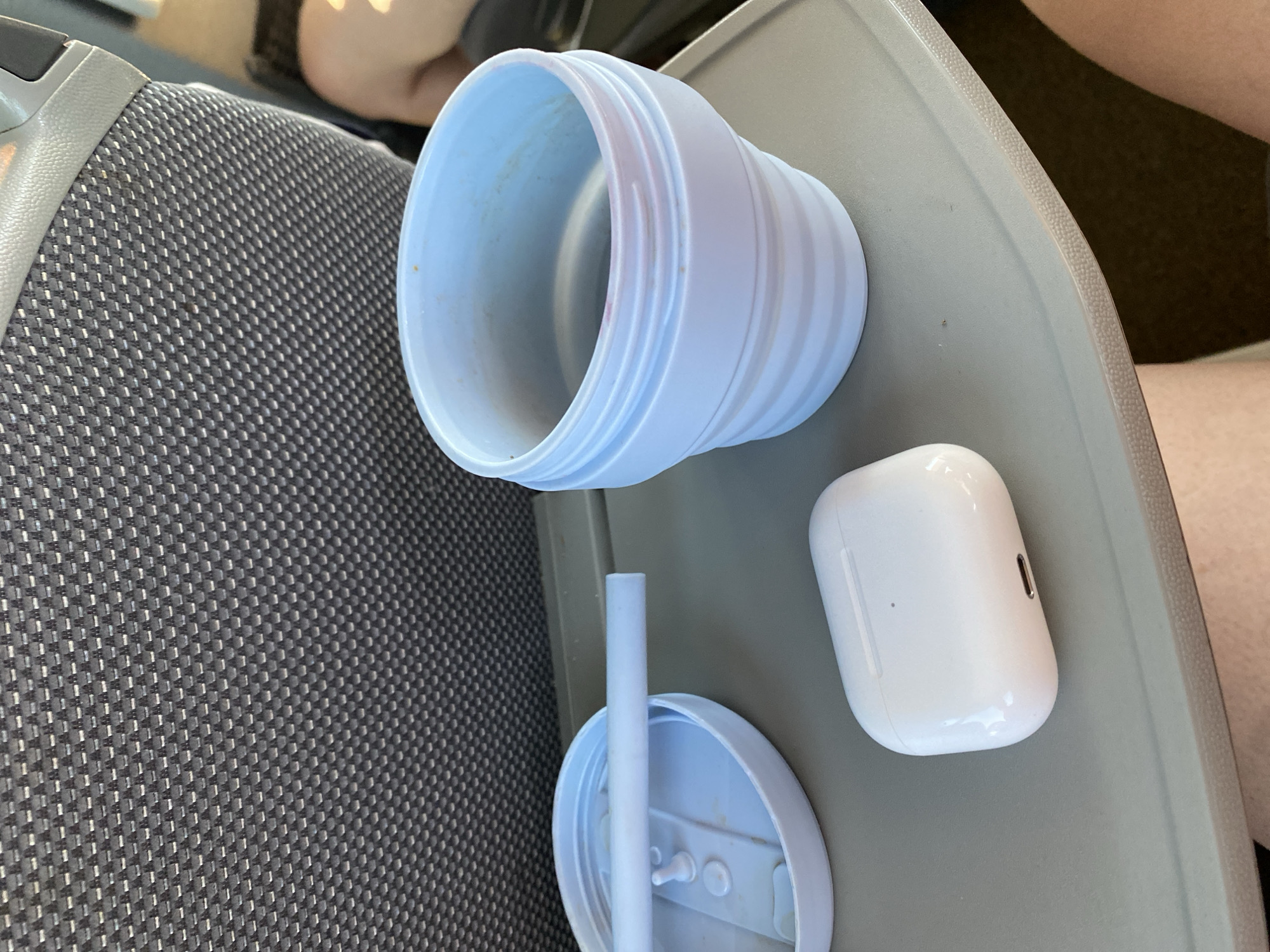A close-up shot captures a grey airplane seat with a tray table holding several items. On the tray, there is a light blue collapsible cup, a light blue lid with a straw inserted, and a white wireless earbud charging case. The background shows the textured fabric of the airplane seat and a person's bare legs. The scene appears to be set on a flight, likely in Kápolnásnyék, Hungary, though this location is not directly verifiable from the image itself.