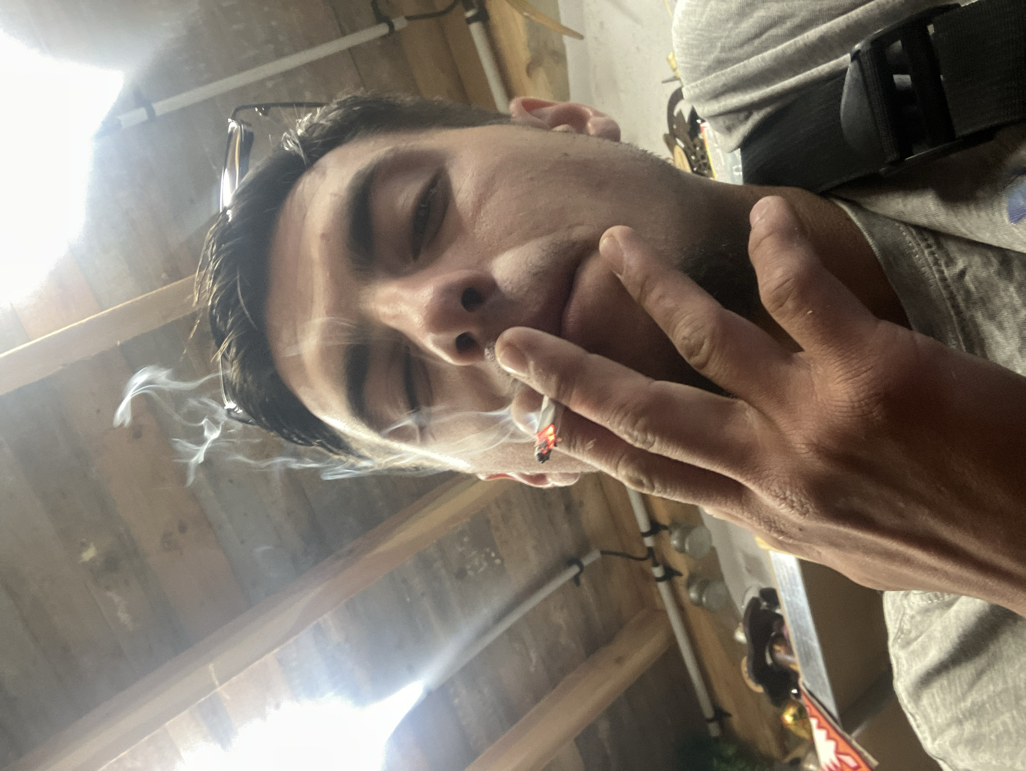 A close-up, angled selfie depicts a man with short dark hair and light skin. He is holding a lit cigarette between his fingers, which are close to his mouth. Smoke rises from the cigarette. The man wears glasses on his head and a grey t-shirt. A black strap is visible over his shoulder. The background appears to be an unfinished interior space with exposed wooden beams and pipes. The location in the city of Komuniga, Bulgaria cannot be confirmed.