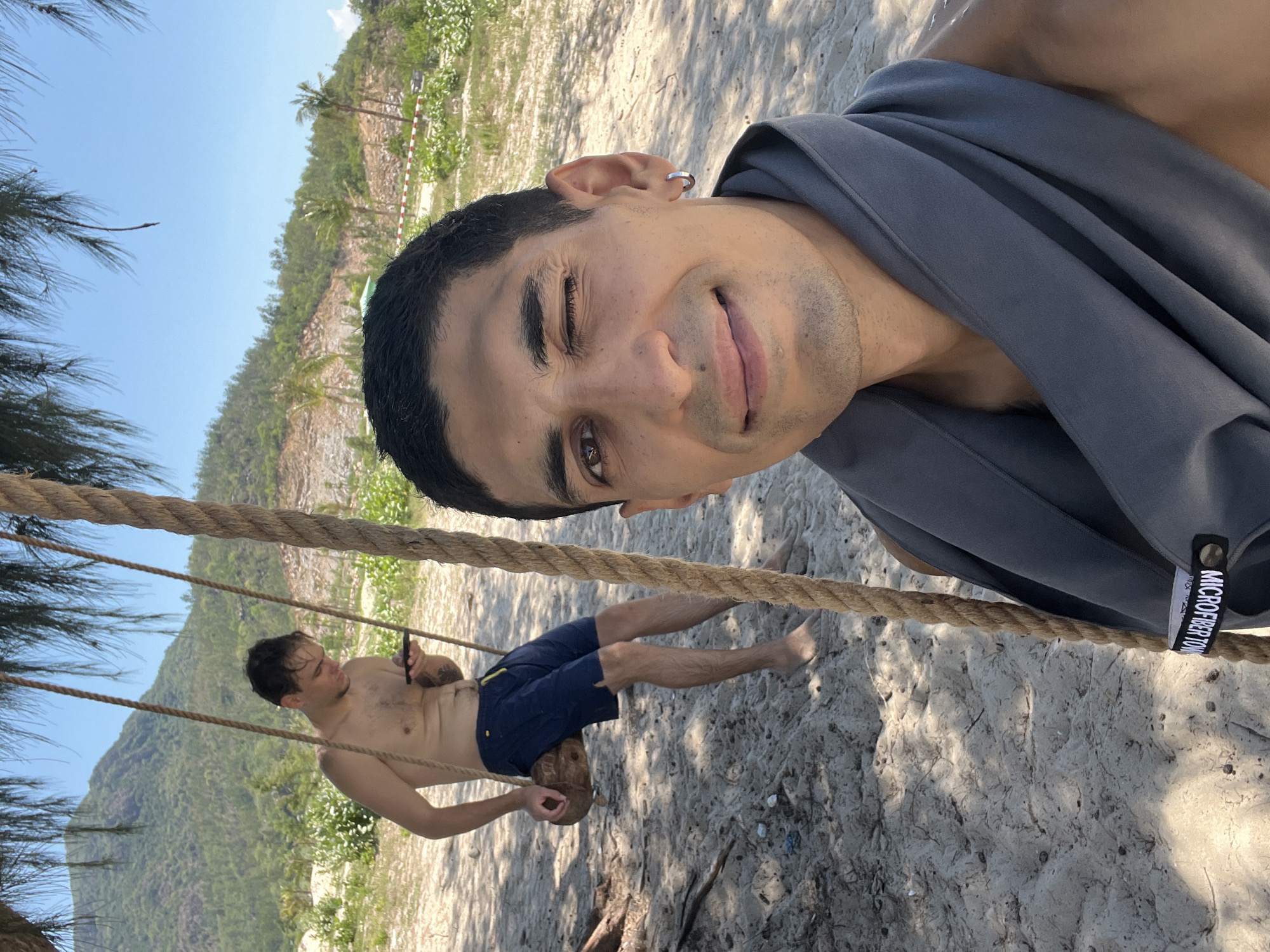 This is a selfie taken on a sandy beach with a lush green hillside in the background. The foreground features a man winking and smiling at the camera, with a grey towel draped around his neck. A thick rope swing hangs from a tree branch, obscuring part of the frame. In the background, another man is sitting on the rope swing, looking at his phone. The environment appears to be a tropical or subtropical location. The presence of the lush hillside suggests a coastal area with significant vegetation. This scene is consistent with the natural beauty of Cong Hai, Vietnam, known for its scenic beaches and coastal landscapes.