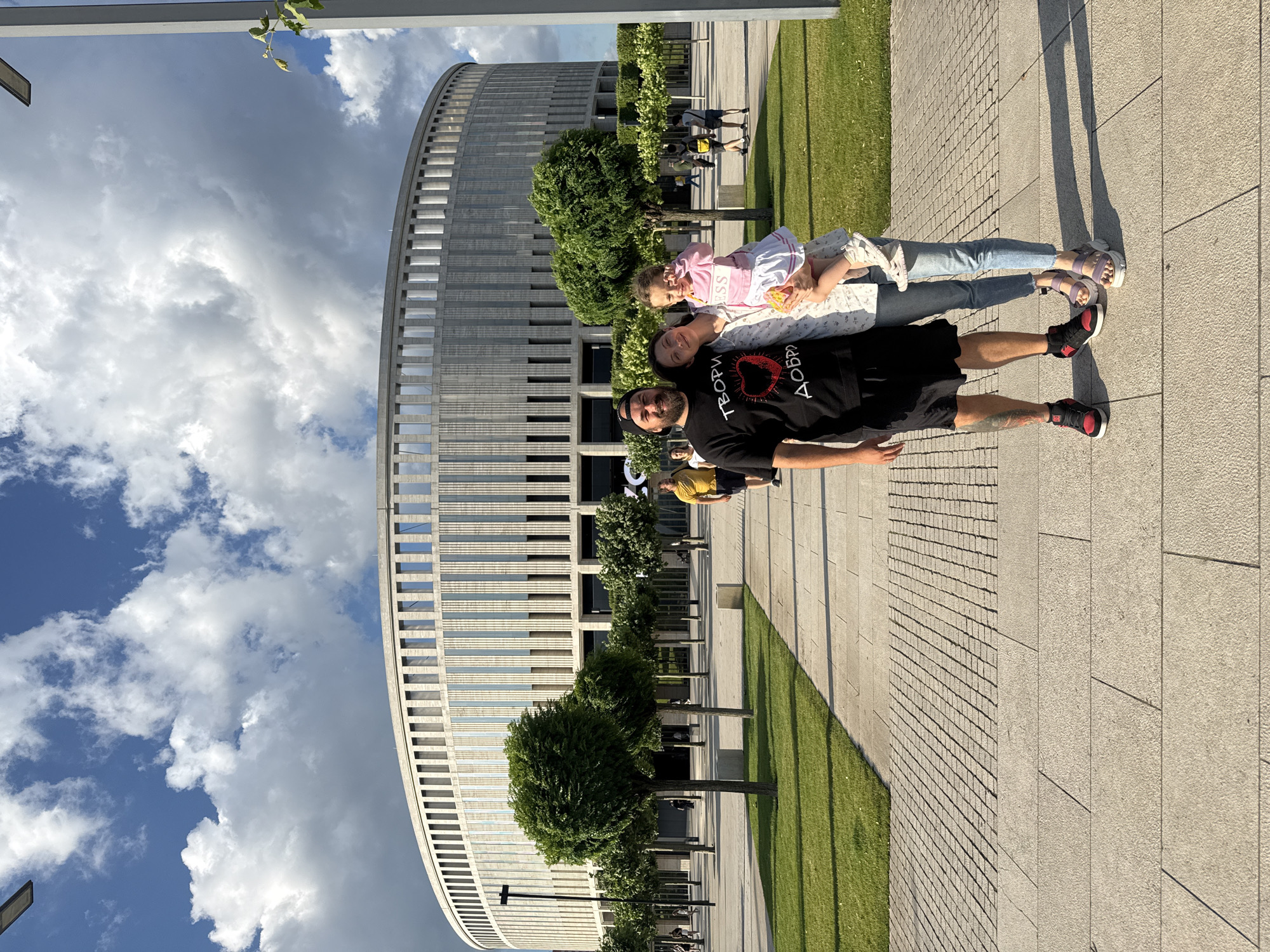 A family of three, a man, a woman, and a young girl, pose for a photo in front of a large, circular, modern building with a textured facade and numerous vertical windows. The man, on the left, wears a black t-shirt with a graphic design of a heart and Cyrillic text, black shorts, and red and black sneakers. He has tattoos on his left leg. The woman, in the center, is holding the girl, who is on the right. The woman is wearing a light-colored, patterned blouse and blue jeans. The girl is wearing a pink sweater with "BOSS" written on it and a white skirt. She has light-colored shoes. The family is standing on a paved walkway. To their left is a well-maintained lawn and several evenly spaced, rounded trees. In the background, beyond the family and trees, other people are visible walking. The sky above is blue with scattered, fluffy white clouds. The building appears to be a stadium or arena, given its circular shape and scale. The environment suggests a public space, possibly a park or plaza surrounding the building.