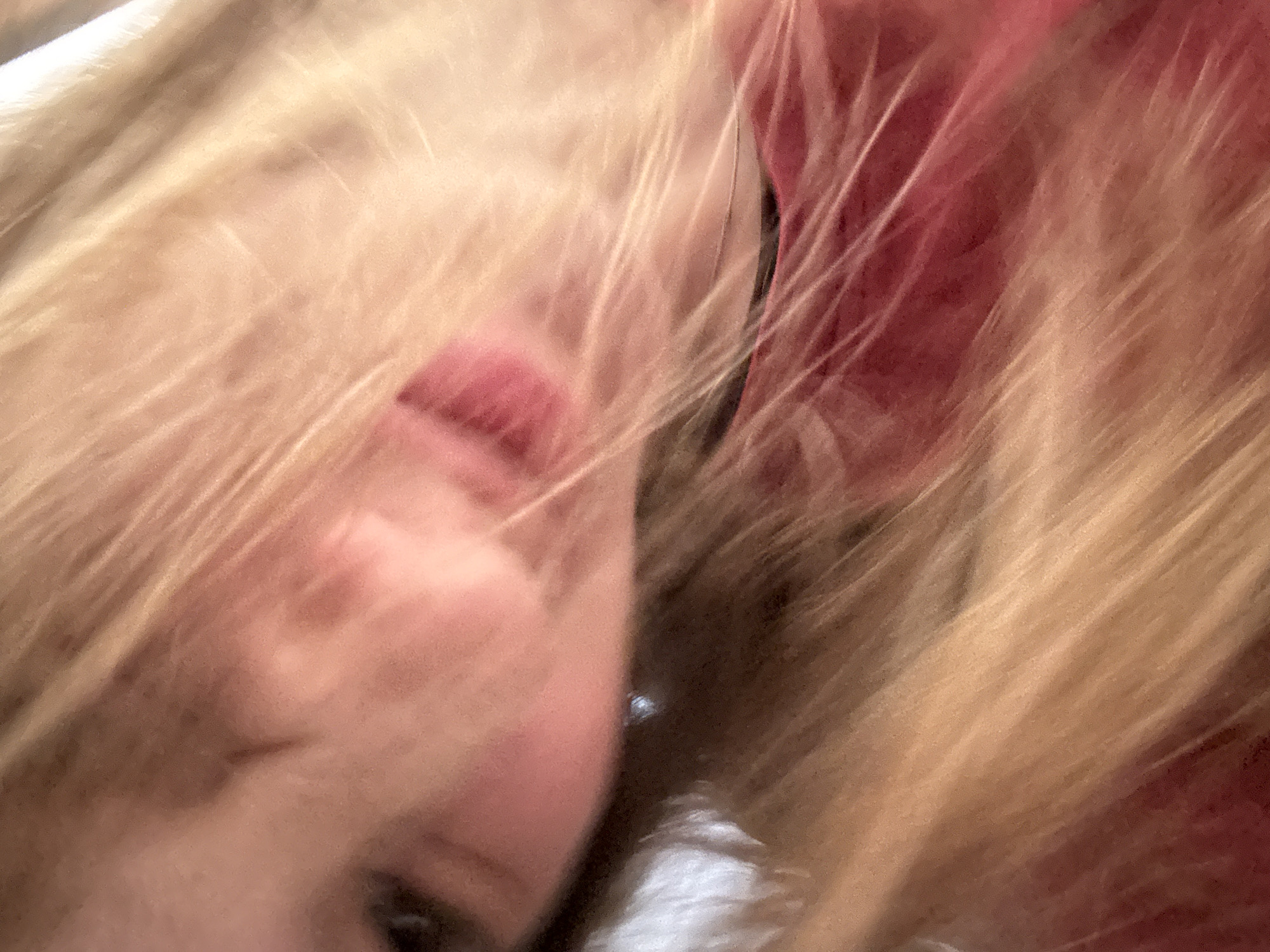 The image is a close-up, slightly out-of-focus shot of a person's face, tilted sideways. The person has fair skin and blonde hair, which is falling across their face, obscuring much of it. They are wearing a red garment, likely a shirt or dress. The lighting is warm and casts soft shadows, creating a gentle and intimate atmosphere. The background is indistinct, suggesting an indoor setting. The exact location within the city of Unknown, Russia, cannot be determined from the visual information provided.