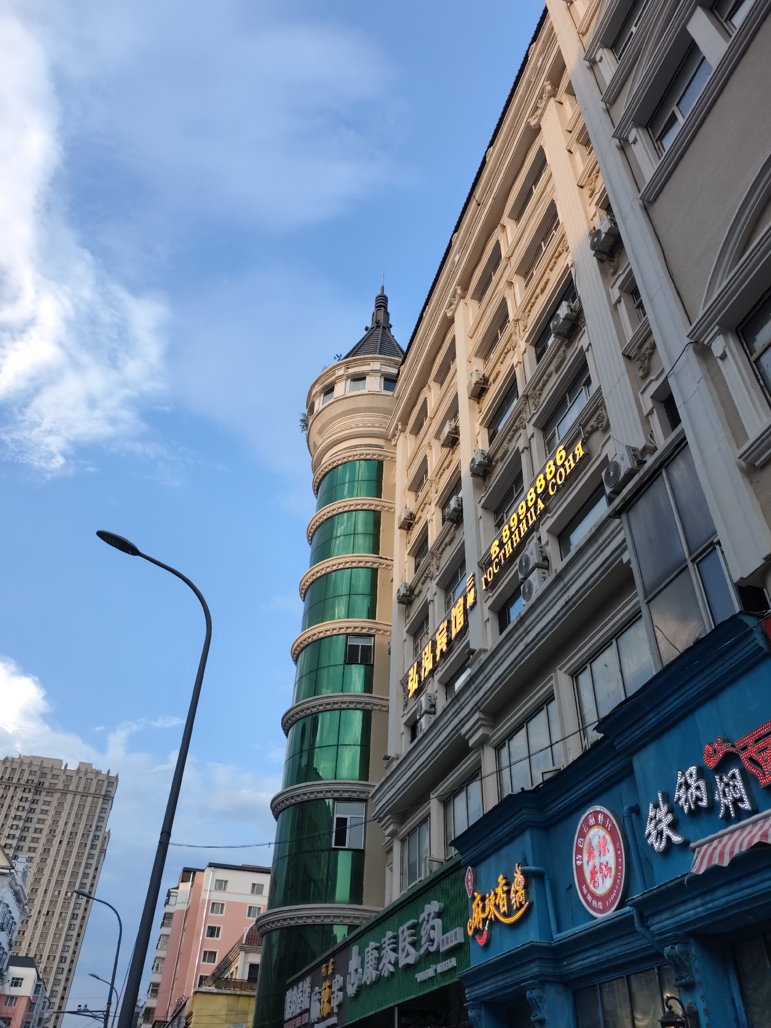 This is a low-angle, wide shot of a city street in Aihui, China, during the day under a partly cloudy sky. The foreground features a bright blue facade with red and white striped awnings and illuminated signs in Chinese characters, some of which appear to be for a restaurant. To the left of this, a green facade with white lettering is partially visible. Behind these, a tall, cream-colored building with multiple floors and air conditioning units is prominent. To the left of the cream building is a distinctive circular tower with green, reflective glass panels and white decorative trim. A dark, curved street lamp extends into the frame from the lower left. Further in the background, a very tall, modern skyscraper with a patterned facade is visible on the left, and a pink and white building with numerous windows is seen to the right of the circular tower. The overall scene depicts a bustling urban environment with commercial establishments and residential buildings.