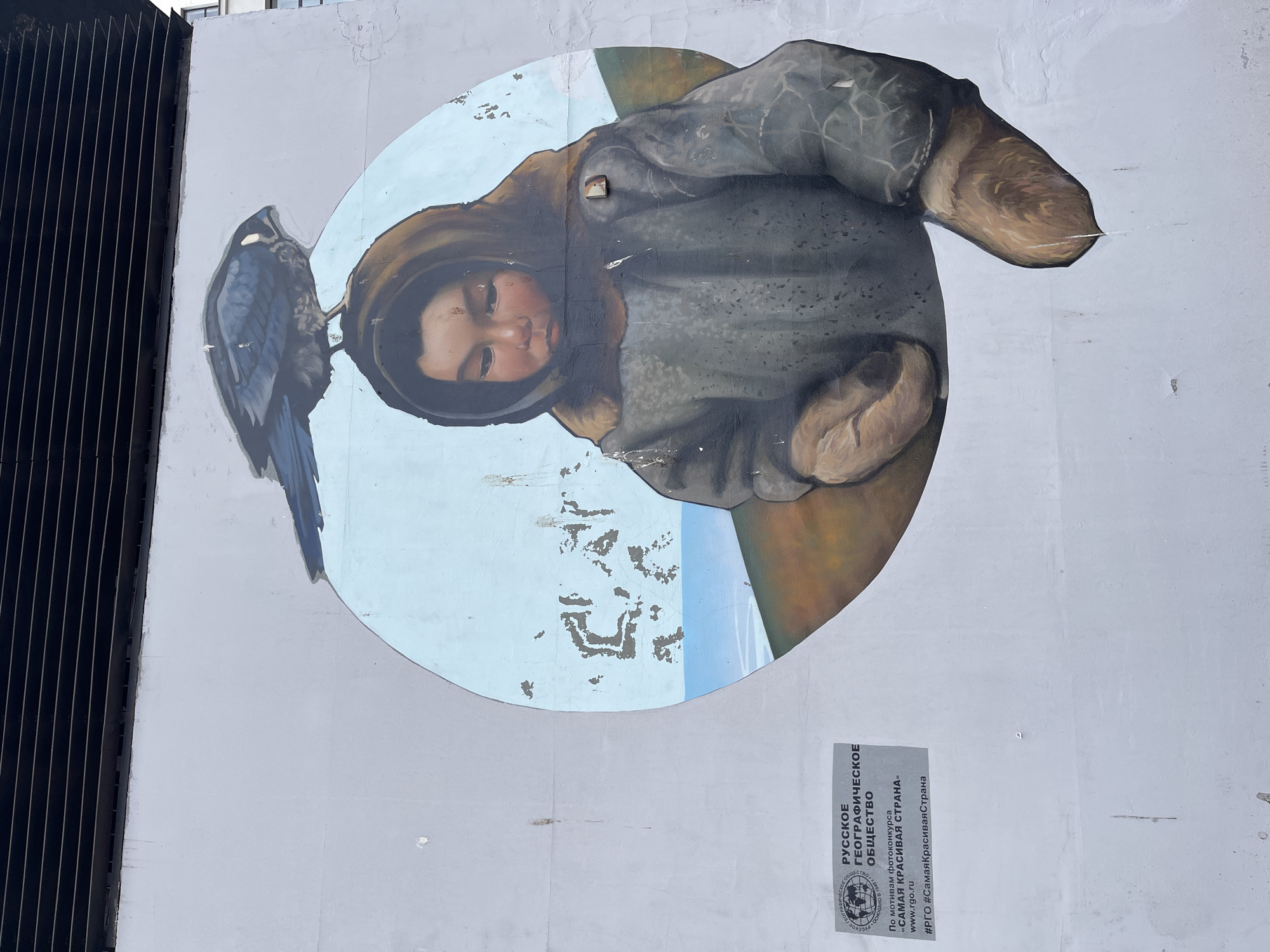 The image shows a mural on a light-gray wall, with a roundel containing a portrait of a person in a hooded coat, looking downward with eyes closed. A blue-gray bird is perched on the left side of the roundel. To the left of the mural is a section of dark vertical bars. To the right, beneath the roundel, is a gray informational plaque with Russian text. The plaque includes the text "РУССКОЕ ГЕОГРАФИЧЕСКОЕ ОБЩЕСТВО," which translates to "Russian Geographical Society." The text also mentions a photo contest called "САМАЯ КРАСИВАЯ СТРАНА" which translates to "The Most Beautiful Country." The location is Moscow, Russia.