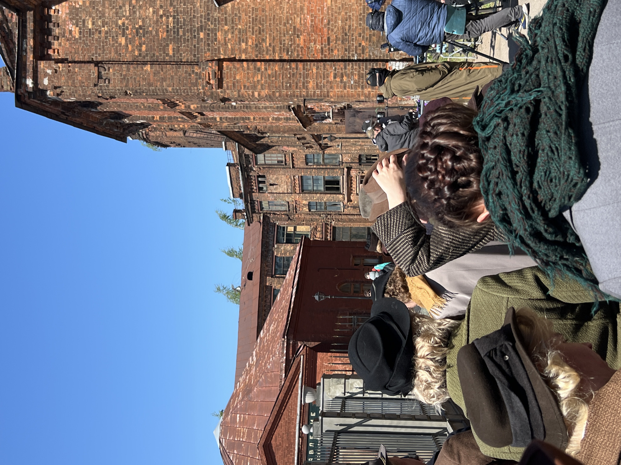 A sideways-oriented medium shot captures a gathering in what appears to be an urban environment, likely Sestroretsk, Russia. The image centers on a crowd of people, their backs and shoulders primarily visible. Several individuals are wearing vintage or period-style clothing, including hats and scarves, suggestive of a theatrical production or historical reenactment. The architecture in the background includes tall brick buildings, indicative of an industrial or historical district. The sky is clear blue, suggesting a sunny day. There are metal railings and what appears to be street furniture or equipment scattered around, possibly indicating a set or staging area. Several people are observing with cameras.