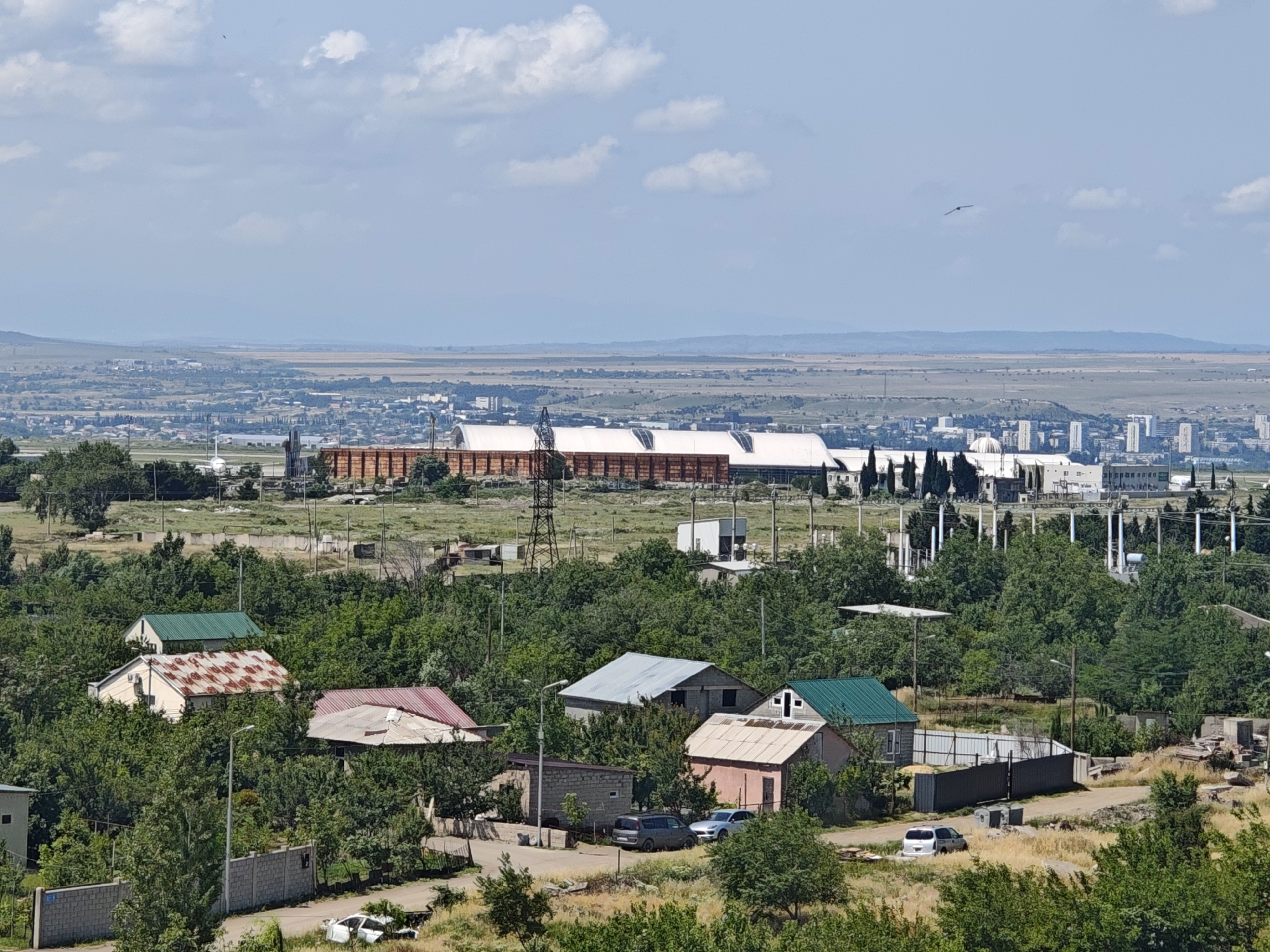 This is an eye-level, wide shot of a residential area with a distant urban landscape and a large industrial building in the mid-ground. In the foreground, several small, modest houses are nestled among lush green trees. Some houses have red or green metal roofs, while one has a rusted, patterned metal roof. A few vehicles, including a dark SUV and a silver sedan, are parked on a dirt road. A grey brick wall runs along the left side of the foreground. In the mid-ground, a long, rectangular industrial building with a white, segmented roof dominates the scene. It is flanked by power lines and pylons. Beyond the industrial building, the city unfolds with various buildings, including high-rises, and a hazy horizon of rolling hills under a partly cloudy sky. A bird is visible in flight in the upper portion of the sky. The overall impression is a mix of rural residential life and industrial development.