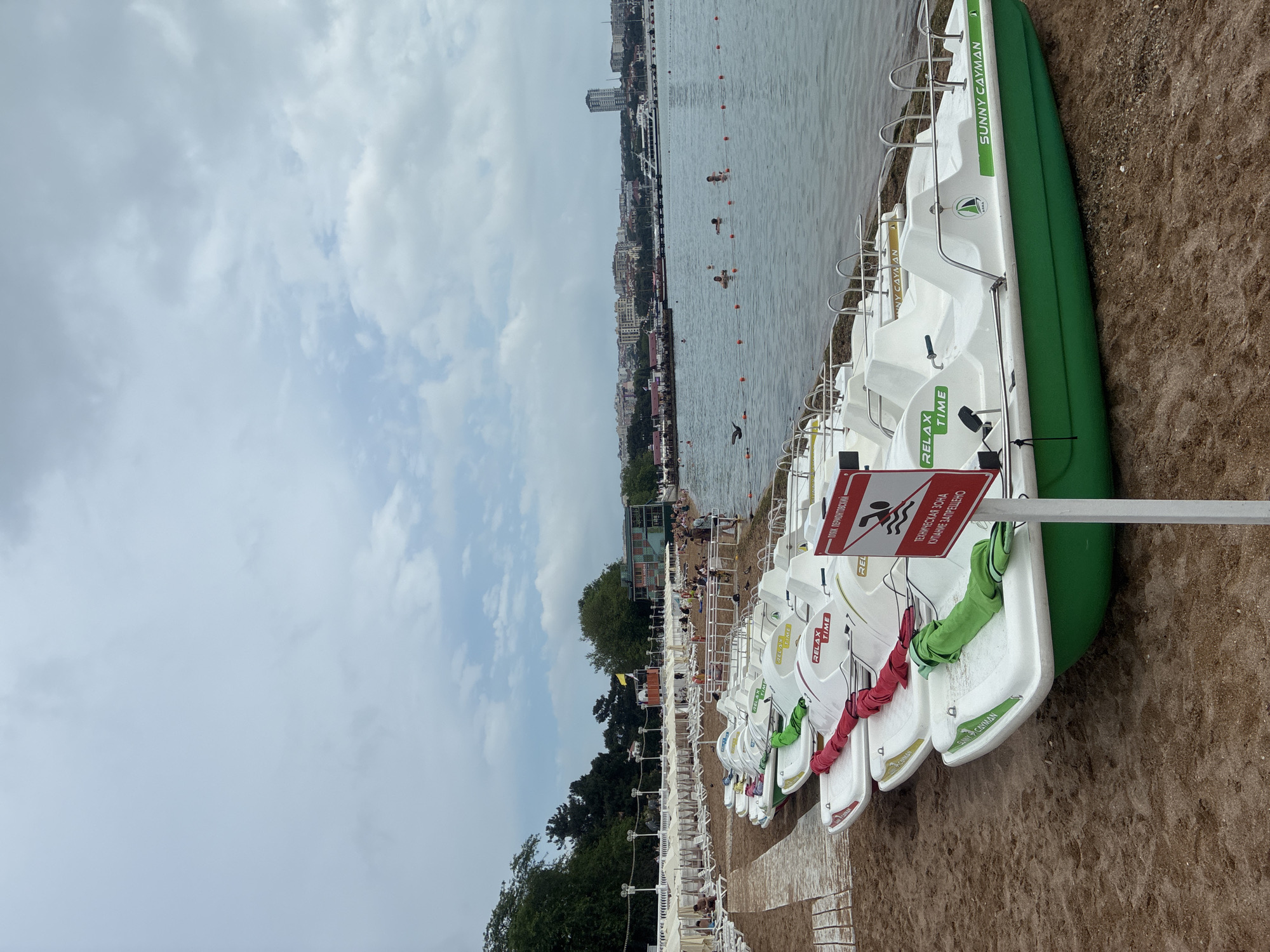 The image captures a sandy beach in Gelendzhik, Russia, with a row of white and green pedal boats lined up on the shore. The sea is calm, with several people swimming and enjoying the water. In the background, a coastal city skyline is visible, featuring buildings and a pier. A red and white sign with a prohibition symbol and text is placed near the pedal boats. The sky is overcast with scattered clouds.