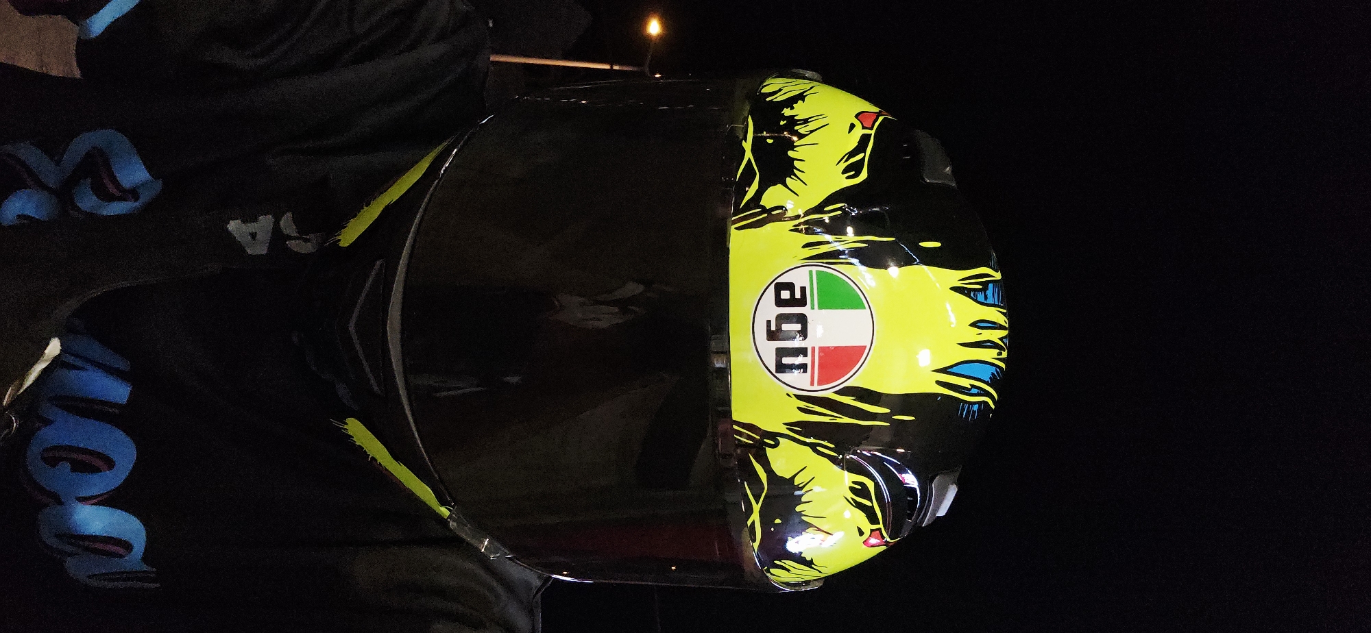 A motorcycle helmet with an AGV logo is positioned against a dark background. The helmet features a yellow and black design with accents of green, red, and blue. The logo is prominently displayed on the side of the helmet. The surroundings appear dark, and the location and date are unknown.