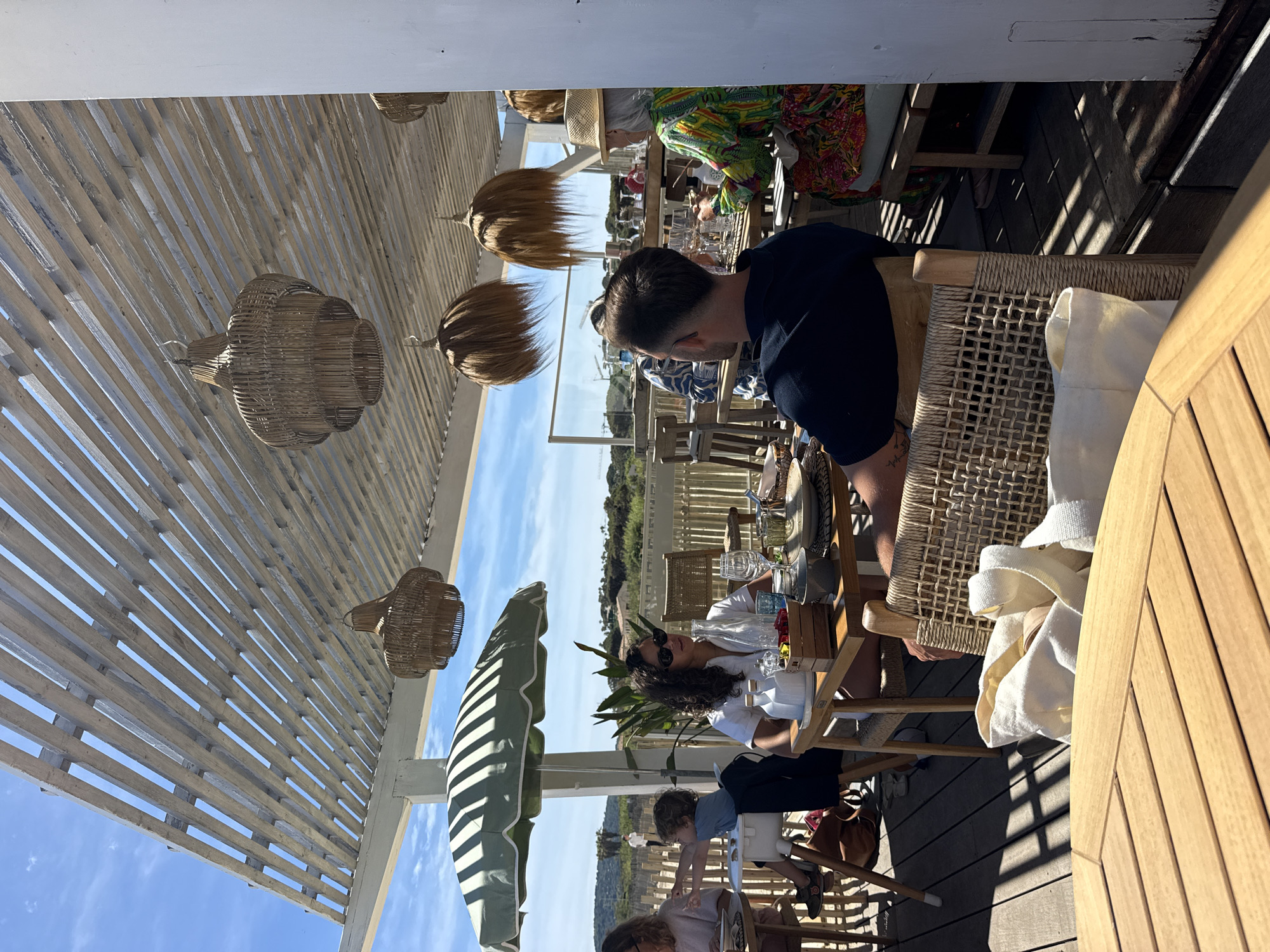 The image depicts an outdoor dining scene at what appears to be a restaurant or bar in Ramatuelle, France, based on the coastal vegetation in the background and the architectural style. Several people are seated at wooden tables, with some individuals in the foreground and others further back. The diners are engaged in conversation or enjoying their meals. A pergola structure with wooden slats covers the area, providing shade. Decorative straw light fixtures hang from the pergola. The flooring is wooden decking. Green and white striped umbrellas provide additional shade. The scene suggests a relaxed, sunlit, and upscale dining experience.