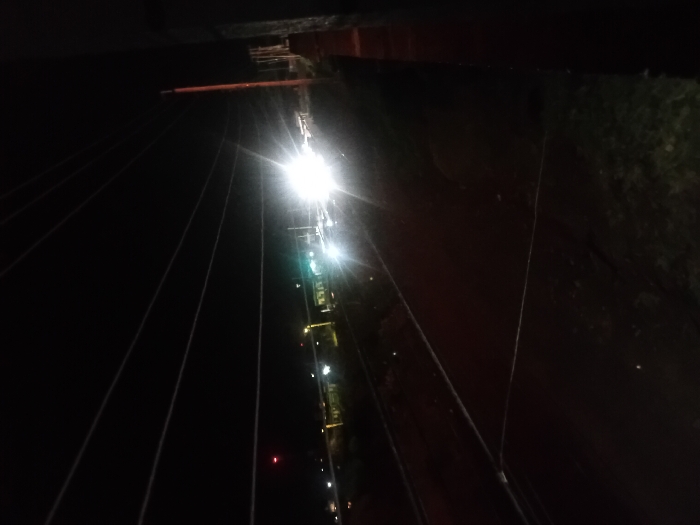 A nighttime view of a suspension bridge's structure. Bright lights illuminate the bridge's tower and cables stretching diagonally across the dark scene. A reddish-brown structure is visible on the right, contrasting with the overall darkness. A series of smaller lights line the bridge's edge. Vegetation can be seen dimly behind the reddish-brown structure on the right, adding depth to the scene.