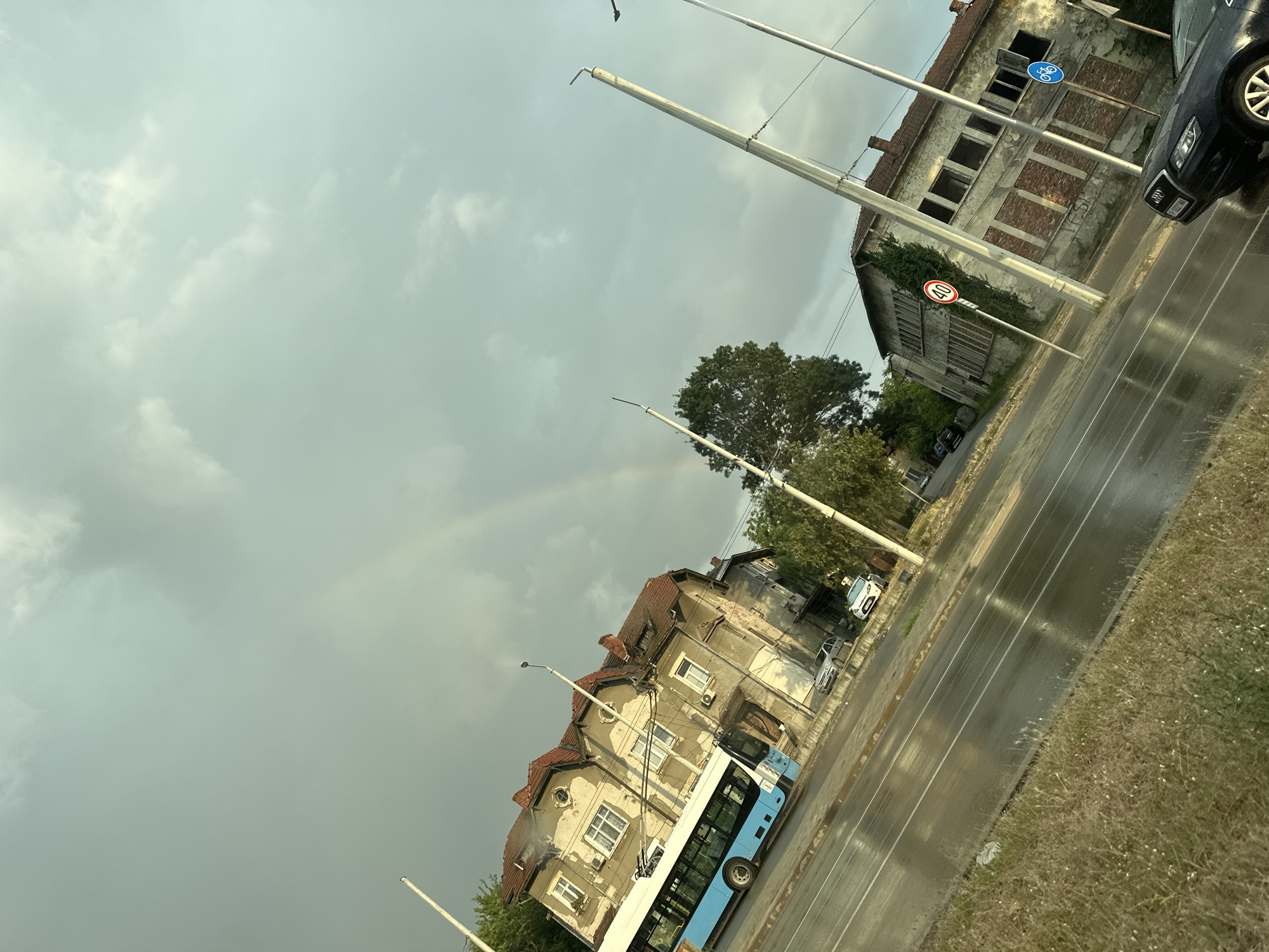 The image depicts a street scene in Ruse, Bulgaria, under a cloudy sky with a faint rainbow visible. The road is wet, suggesting recent rain. A light blue and white trolleybus is the central focus, positioned on the right side of the road and angled towards the viewer.
To the left of the trolleybus are several older buildings. The closest one has a red-tiled roof and light-colored walls with multiple windows. Further down the street, another building appears more dilapidated, with boarded-up windows and exposed brickwork. Streetlights stand on either side of the road.
A black car is parked on the right side of the road, in front of the dilapidated building. Various other cars are visible parked along the street and in front of buildings. Vegetation, including trees and grassy areas, lines the side of the road.
Two traffic signs are visible. One is a circular sign with a red border and the number "40" inside, indicating a speed limit. Another sign features a blue circle with a white bicycle symbol, signifying a cycling path or zone. A third sign, partially obscured by a streetlight, is circular with a red border and a white horizontal bar, indicating a "no entry" or "closed to all vehicles" restriction.
The overall impression is of a somewhat weathered urban environment, possibly on the outskirts of the city, with the lingering effects of rain and an overcast sky. The time of day appears to be late afternoon or early evening, given the lighting and the presence of the trolleybus. No people are clearly visible in the image.