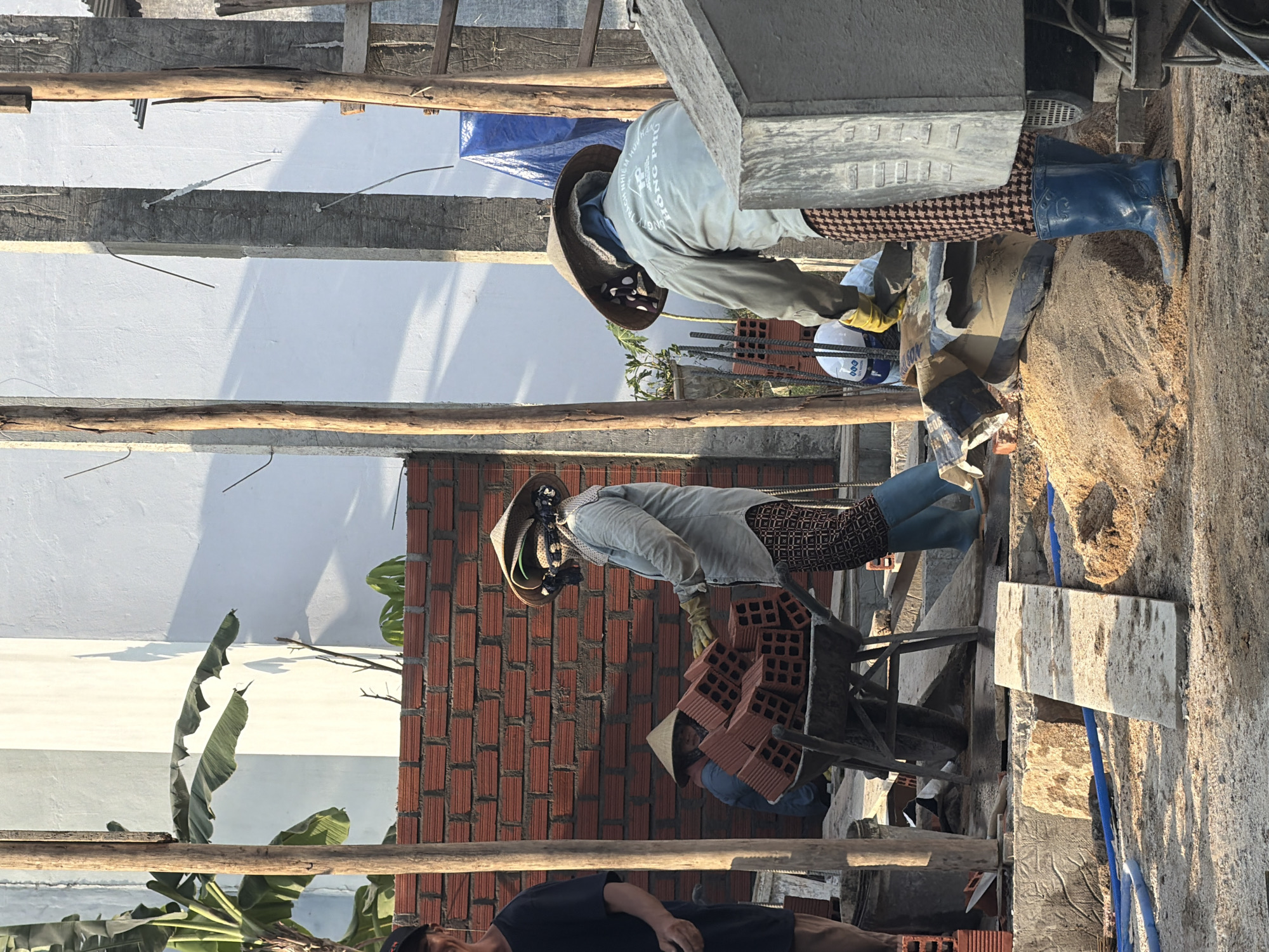 In the city of South Quy Nhon Ward, Vietnam, a construction scene unfolds. Three individuals are present. On the left, a person is partially visible, wearing a black t-shirt and shorts, with a portion of their face in view. In the center, a worker wearing a conical hat and a light-colored shirt is pushing a wheelbarrow laden with red bricks. To their right, another worker, also with a conical hat, is seen from the back, wearing a light-colored long-sleeved shirt and patterned pants, and blue rubber boots. This worker appears to be interacting with the wheelbarrow. The environment is dusty and sandy, with piles of sand, construction materials such as bricks, and wooden support beams visible. A partially constructed red brick wall stands in the background, along with a white wall and some foliage. The lighting suggests it is daytime, possibly late afternoon.