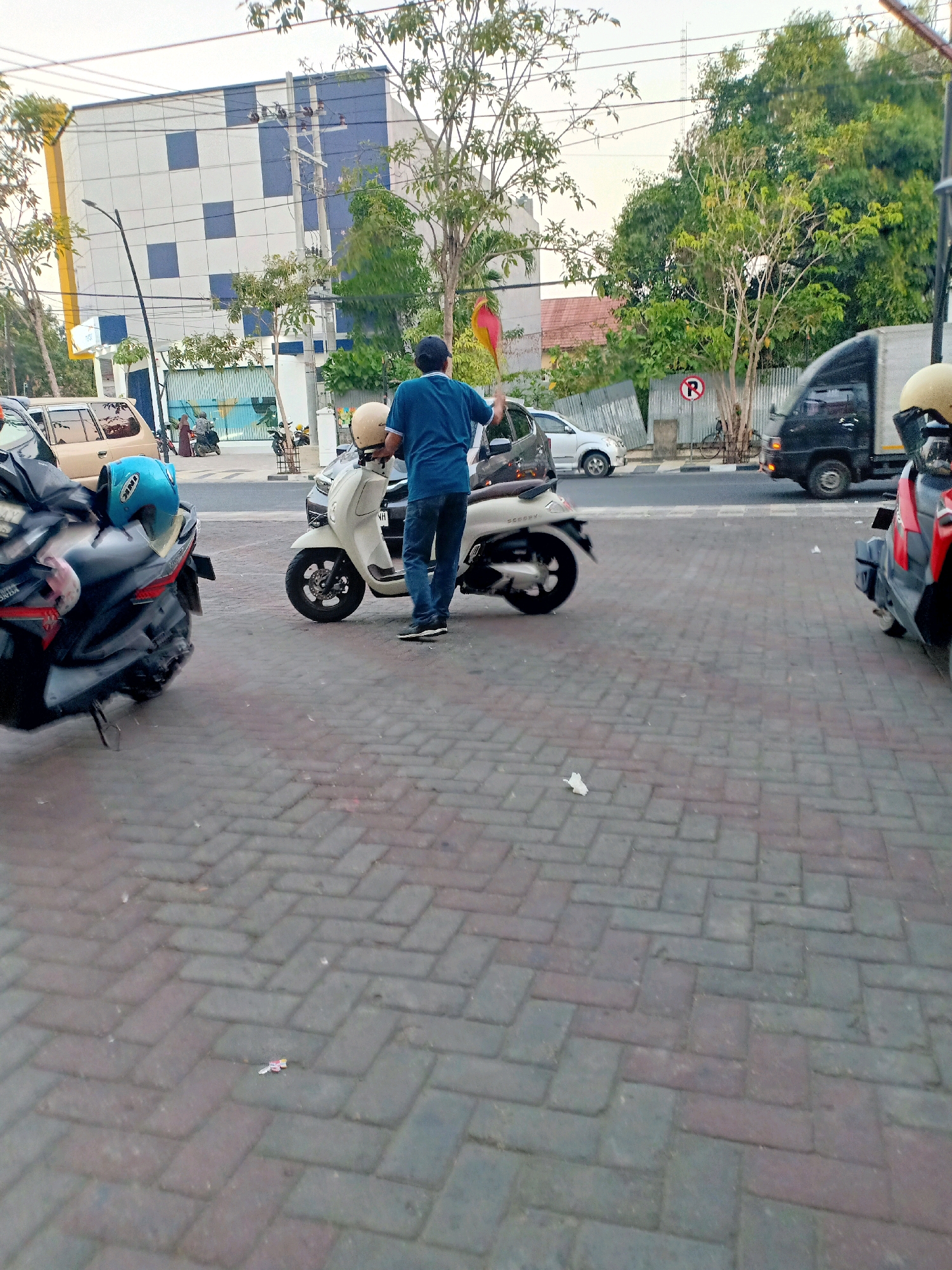 The image depicts a street scene in Bojonegoro, Indonesia, with a focus on parked motorcycles and a person interacting with one of them. In the foreground, two motorcycles are visible on a patterned brick sidewalk. The motorcycle on the left is dark colored with a blue helmet on its seat. The motorcycle on the right is a cream-colored scooter, with a person in a blue shirt and jeans standing beside it, holding a beige helmet. The person appears to be preparing to ride.
Behind the motorcycles, a street runs perpendicular to the viewpoint. Several vehicles are present on the street, including a light-colored car and a black truck. In the background, a multi-story building with a distinctive checkered facade of white and blue squares is visible. Adjacent to the building is a row of trees and a corrugated metal fence. Electrical poles and wires are also present, indicating an urban environment. The sky is overcast. The overall impression is a typical street scene in a commercial or residential area of Bojonegoro.