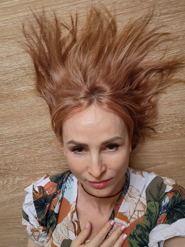An adult female is depicted in a close-up, overhead shot, lying on a light-colored wooden surface with horizontal grain. Her reddish-blonde hair is fanned out above her head. She has fair skin, prominent cheekbones, and wears visible eye makeup and light pink lipstick. She is attired in a collared shirt featuring a tropical pattern in white, orange, green, and brown tones, and a thin red choker-style necklace. Her left hand, with light pink nail polish, is partially visible resting on her upper chest. The subject maintains direct eye contact with the camera. The specified location of Chaikovsky, Russia, cannot be visually confirmed from the image content.