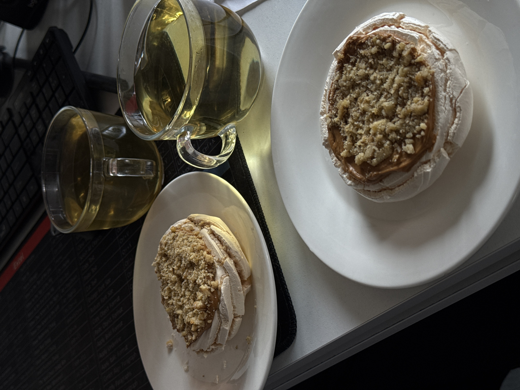 The image presents an overhead view of a tabletop scene featuring refreshments and a work area. Two clear glass mugs, one larger and one smaller, are filled with a translucent, light amber liquid, likely tea. The larger mug is positioned at the top left, while the smaller one is below it.
To the right, two white oval plates each hold a delicate dessert. These desserts appear to be meringue shells, filled with a creamy, light brown filling and generously topped with finely crushed nuts or crumbs. One dessert is prominently displayed in the foreground on its plate, while the other is slightly further back and to the right on a similar plate.
The background on the left side of the image reveals elements of a workspace. A black computer keyboard is partially visible, resting on a black desk mat or mousepad. This mat is inscribed with numerous white text labels, which are identifiable as Microsoft Excel shortcuts and formatting options. Visible text includes "Excel", "Ctrl+C", "Ctrl+V", "Save", "Open", "Print", "Bold", "Italic", "Underline", "Align Left", "Center", "Font Size", and "Conditional Formatting", among others.
The lighting is soft and appears to be indoor, possibly artificial, creating subtle reflections on the glass mugs and plates. The overall scene depicts a quiet break with tea and dessert in what appears to be a home office or office setting, indicated by the computer accessories and work-related text. No people are visible, and the time of day is not explicitly discernable, though the lighting suggests an indoor setting rather than bright daylight. The location context is Yerevan, Armenia.