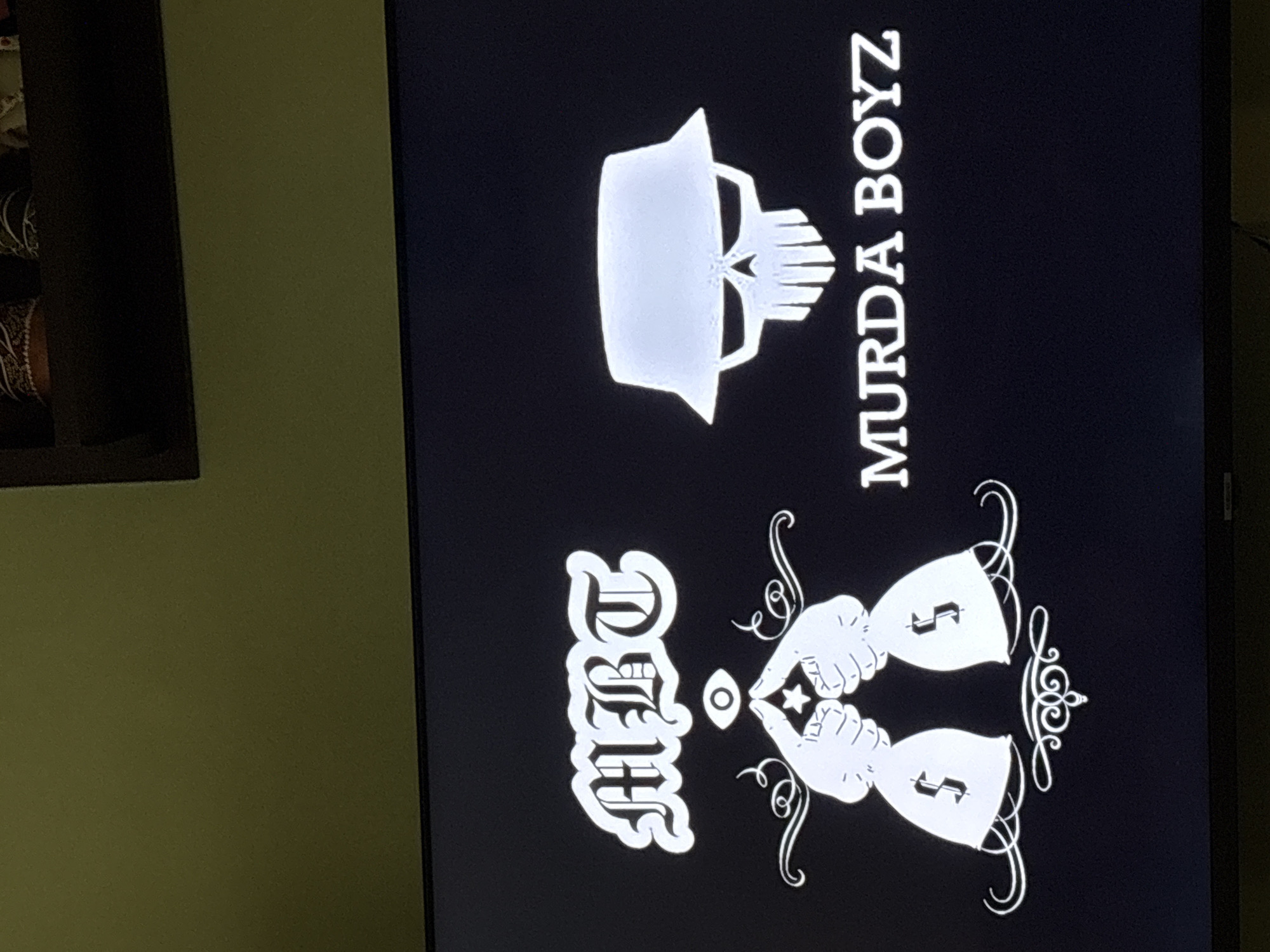 The image shows a black screen displaying white graphics. At the top is a skull wearing a bucket hat, next to the text "MURDA BOYZ" oriented vertically. Below is the text "DRFT" with two hands pointing towards an oval shape. Beneath the hands are two objects resembling chalices with dollar signs on them, surrounded by ornate decorative swirls. The image appears to be a promotional image or logo. There is no evidence in the photo to indicate a location in Plovdiv, Bulgaria.