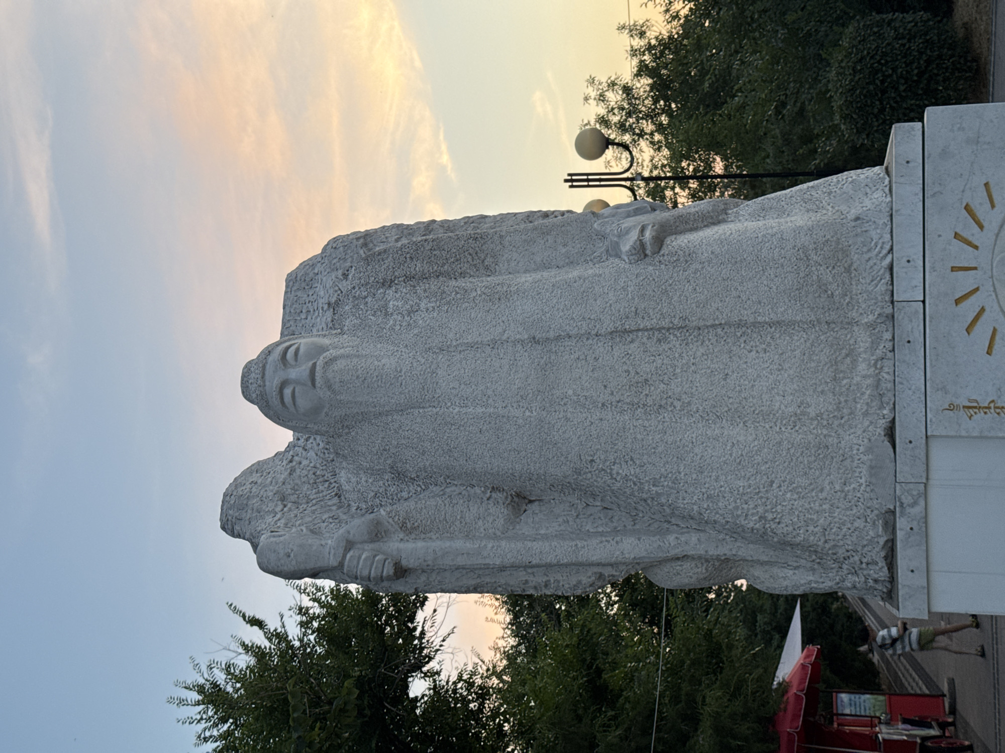 A large, rough-hewn stone statue of an ancient figure stands prominently in Elista, Russia. The statue, depicting a person with a long beard and wearing robes, is carved from light-colored stone with a textured surface. The figure holds a staff in its left hand. It is positioned on a white marble pedestal that has decorative golden symbols resembling rays of a sun.
The statue is outdoors, set against a backdrop of lush green trees. In the distance, a street lamp with two globes is visible, and the sky above shows a gradient of soft blues and oranges, suggesting either sunrise or sunset. To the left of the statue, a red food cart with a white umbrella canopy is partially visible, and a person wearing a striped shirt and shorts is walking away from the cart, towards the statue. The overall environment appears to be a park or public space.