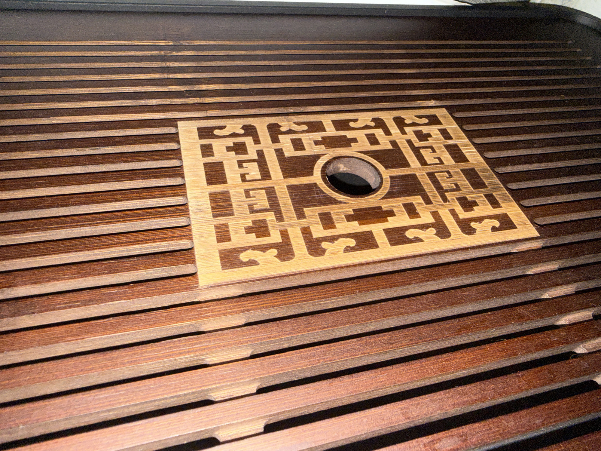 The image is a close-up, angled shot of a wooden surface. The surface is composed of vertical, narrow wooden slats with a dark brown hue, varying slightly in shade and exhibiting a visible wood grain. Overlaid on this slatted surface, towards the center, is a square panel made of a lighter wood, possibly bamboo, featuring an intricate geometric pattern. This pattern has cut-out sections and includes a circular hole in its center. The lighting creates shadows between the slats, emphasizing the texture of the wood. The image does not depict any recognizable landmarks or features that would indicate a specific location in Reppolovo, Russia, or any other city.