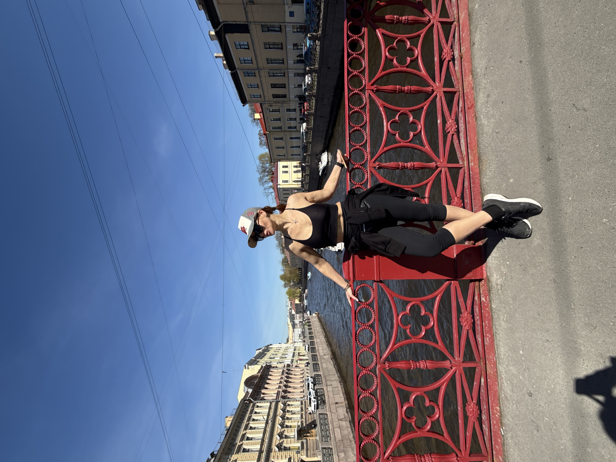A full-length shot shows a person on a bridge. The bridge has a red latticework railing. The person, presumably female, is wearing a black tank top, black leggings, and sneakers. They are also wearing a baseball cap and sunglasses. The person is resting their arm on the railing. Beyond the railing is a body of water and the buildings of Saint Petersburg. The sky is blue with some wires visible. The image is taken at an angle, with the horizon line running vertically. The location is likely Saint Petersburg, Russia, given the style of architecture.