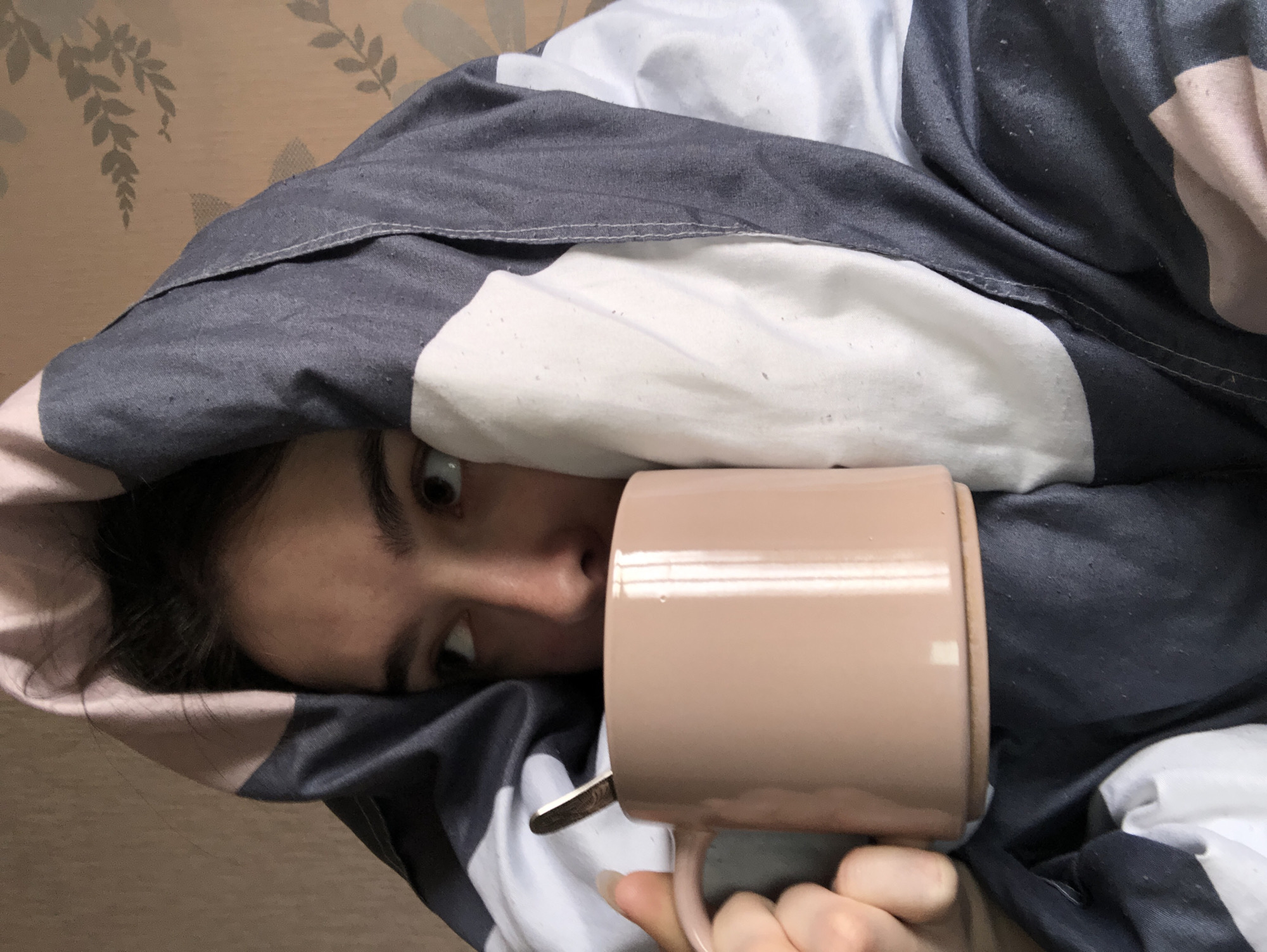 A woman lies in bed, partially covered by a grey, white, and pink duvet. Her eyes are visible, looking slightly downward. A light brown ceramic mug with a spoon inside is held up to her face. The background appears to be a light brown wall. The location of the image is presumed to be in Chaikovsky, Russia, due to the instructions provided.