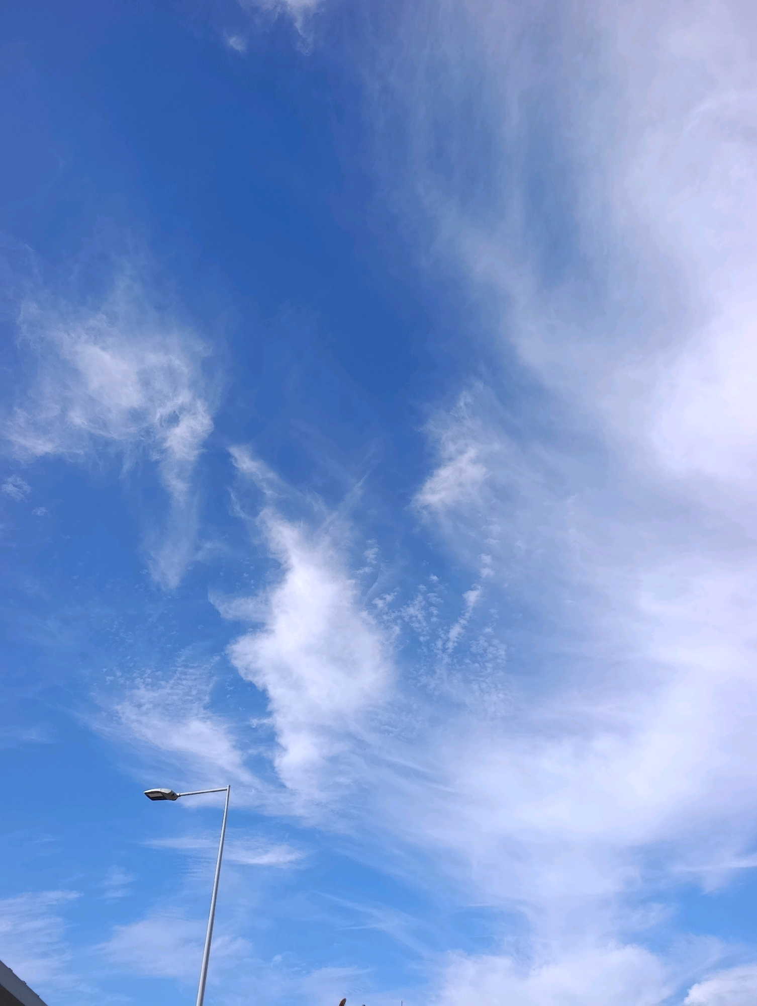 The image depicts a bright, daytime sky filled with wispy white clouds against a vivid blue backdrop. A modern, grey street lamp with a rectangular fixture stands tall on the right side of the frame. A portion of a dark structure, possibly a building or rooftop, is visible at the bottom left corner of the image, suggesting an urban setting. The perspective is looking upwards, emphasizing the vastness of the sky.