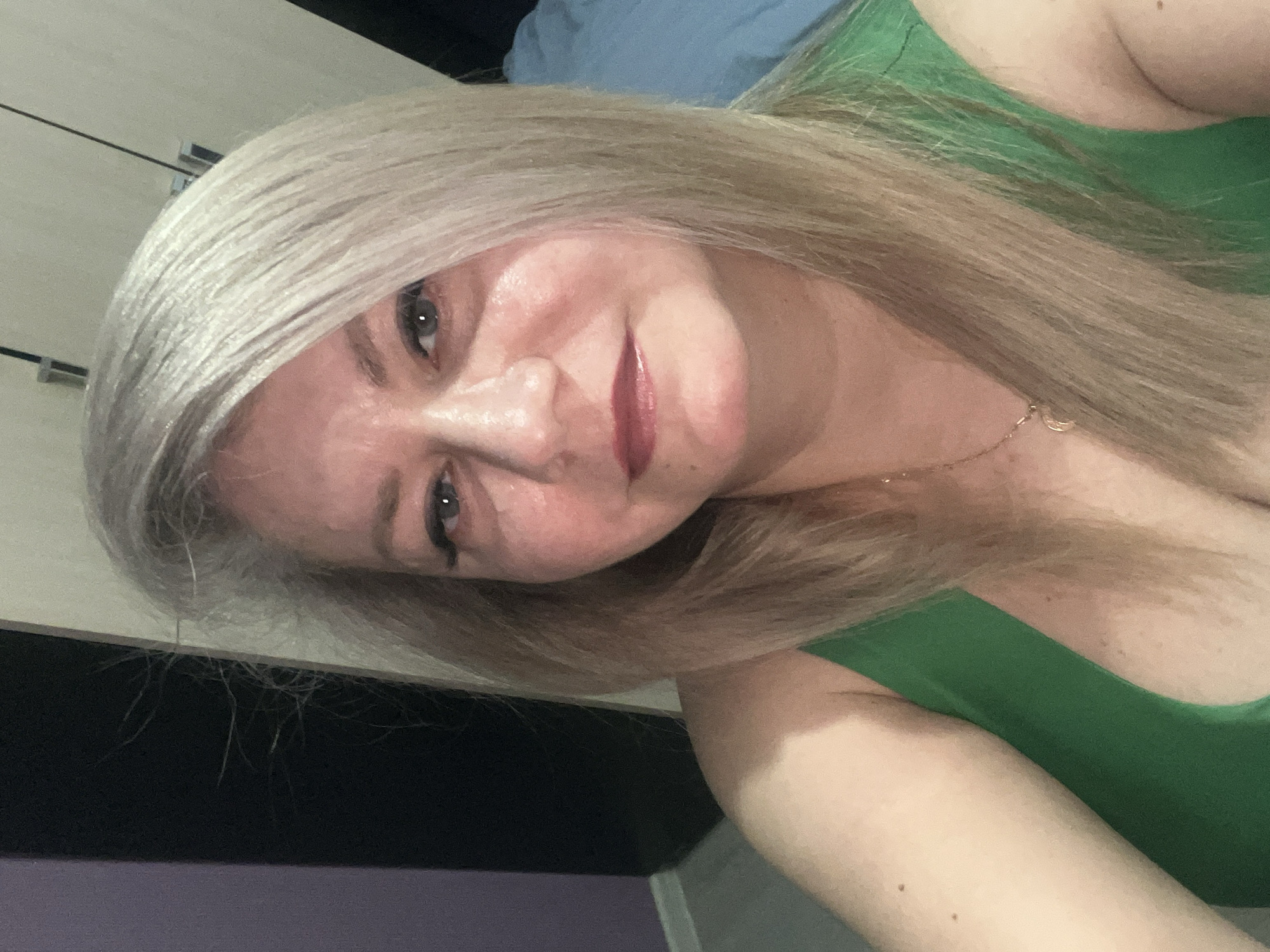 A woman with long, platinum blonde hair is pictured in a selfie. She is wearing a green top. A gold necklace is visible around her neck. In the background, there is a light-colored closet with silver hardware. The location is not discernible from the image. No actions or interactions are visible.