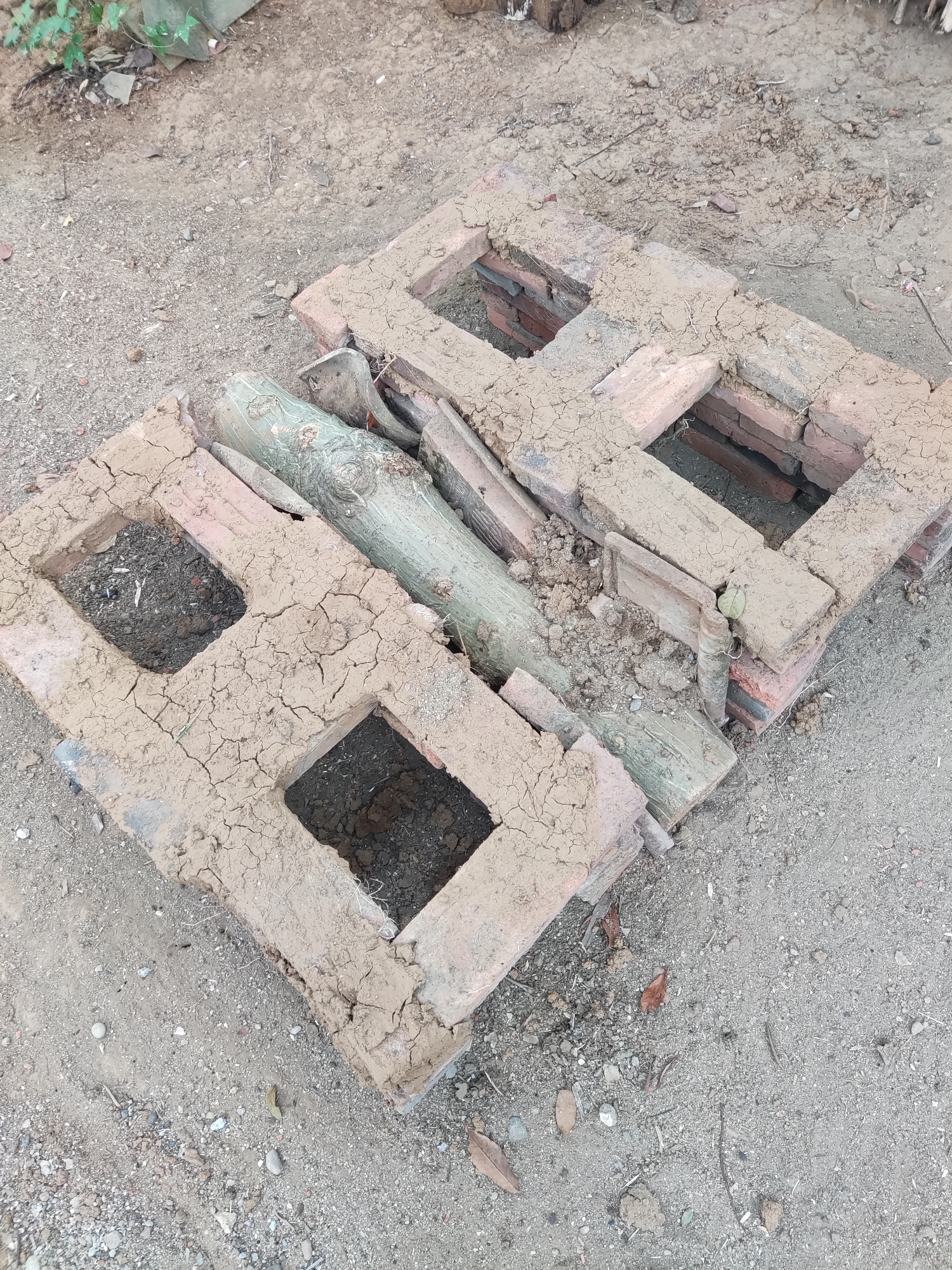 In Ngentrong, Indonesia, two roughly constructed ovens, made of stacked red bricks and covered in dried mud, sit on sandy ground. Each oven has two square openings leading to darker interiors. A few wooden logs, one significantly larger and green, are placed between and on top of the brick structures. The surrounding environment is dry and dusty, with scattered small stones and dried leaves.
